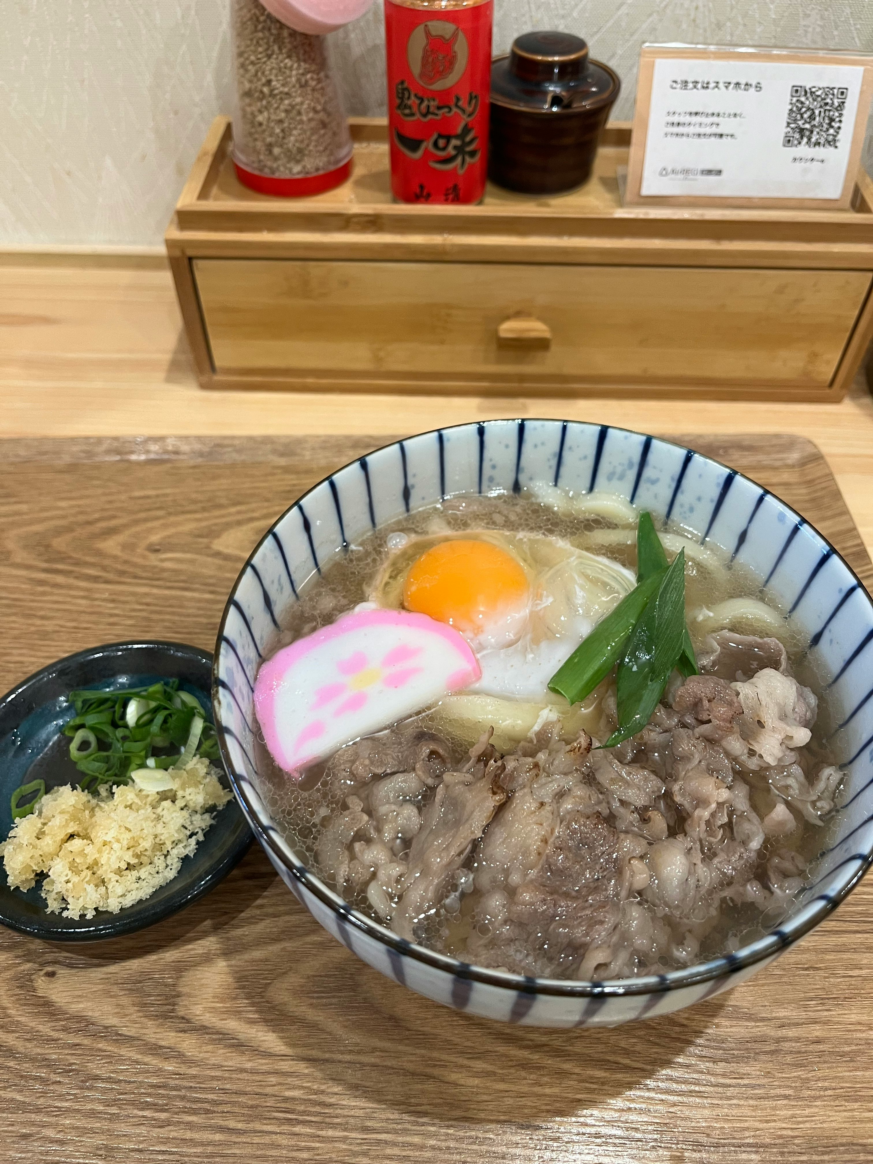 木のカウンターに並ぶ牛すじ煮込みうどんの丼と薬味