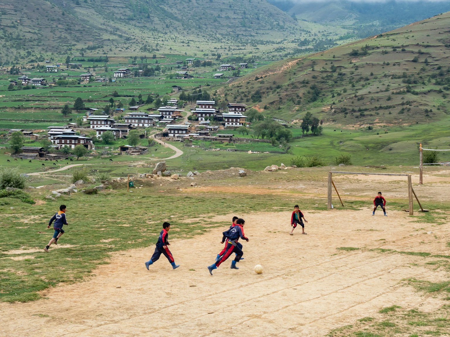 Bambini che giocano a calcio su un campo rurale con montagne e un villaggio sullo sfondo