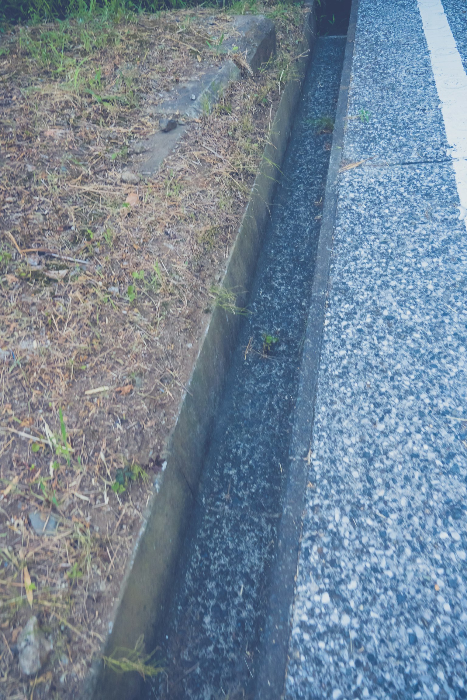 Saluran pinggir jalan dengan kerikil dan rumput di sekitarnya
