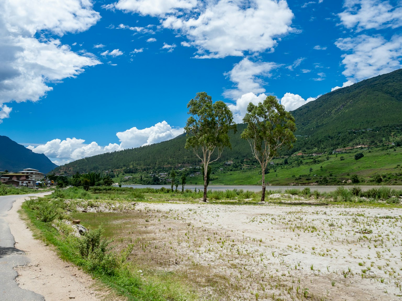 Scenic landscape with blue sky and white clouds Green mountains and river visible Gravel path with scattered trees