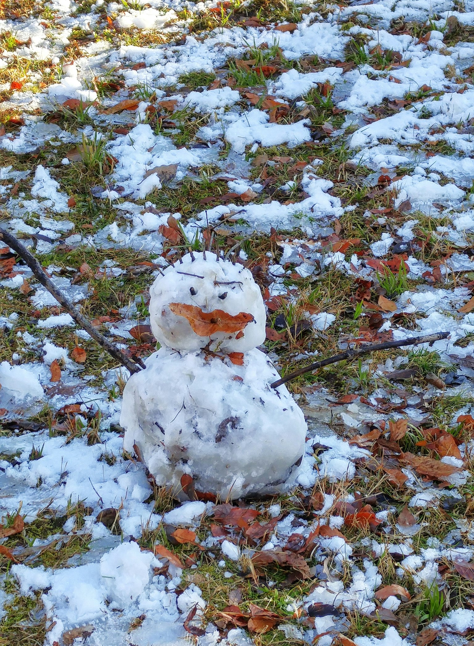 A snowman standing on grass surrounded by snow and fallen leaves