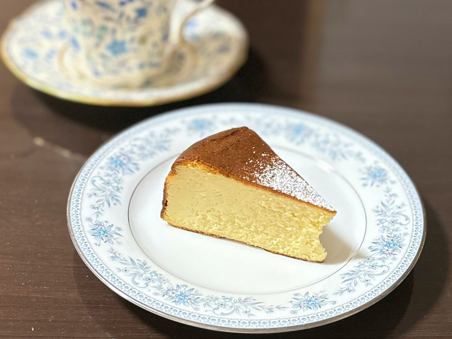 A slice of sponge cake on a decorative floral plate with a cup in the background
