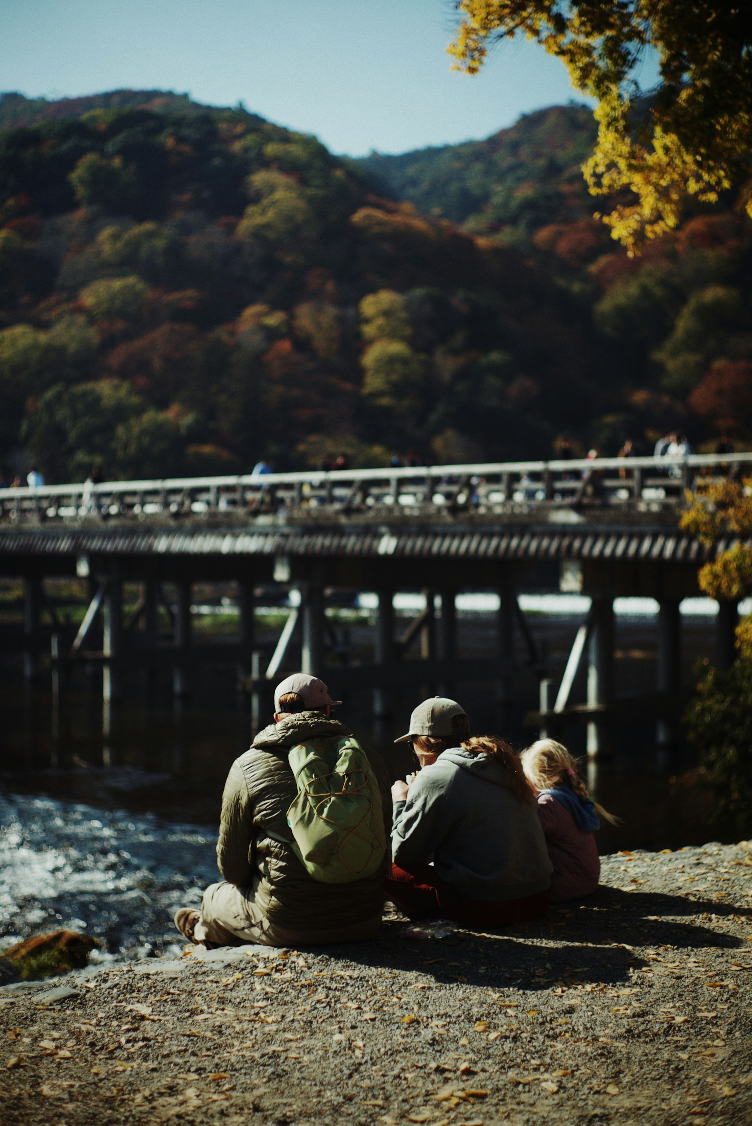 Family sitting by the river with a bridge and colorful mountains in the background
