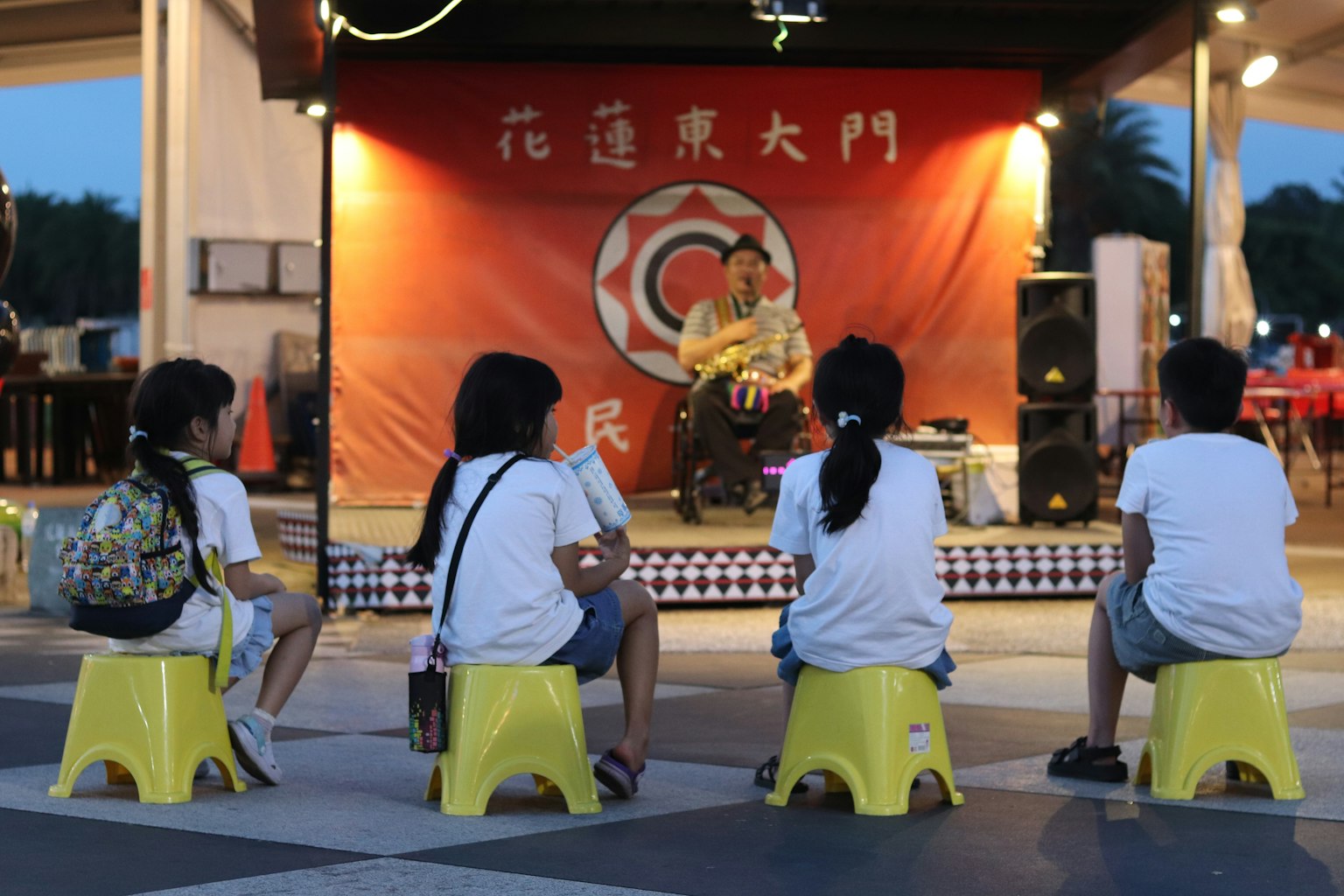 Children sitting on yellow stools watching a performance at night