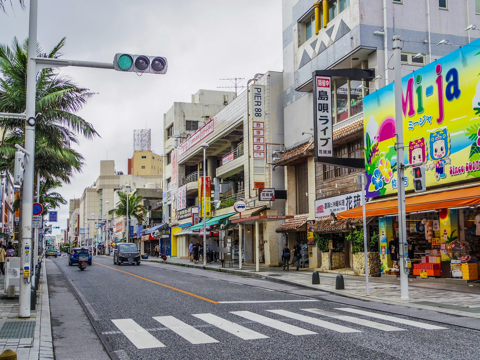 Pemandangan jalan di area perbelanjaan di Okinawa dengan beberapa toko dan jalur pejalan kaki serta pohon palem dan lampu lalu lintas