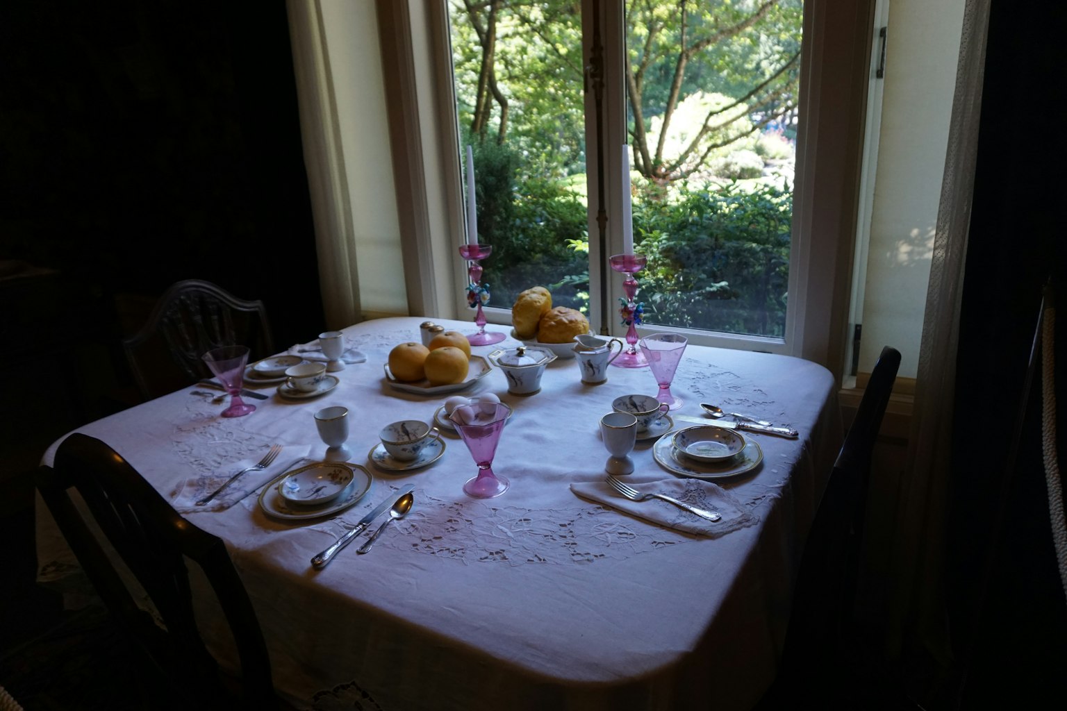 Ein Tischgedeck mit rosa Gläsern und weißen Tellern vor einer grünen Kulisse draußen am Fenster
