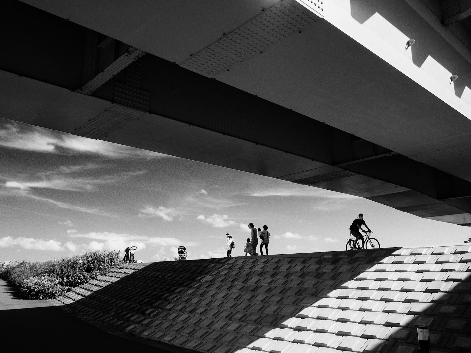 Escena en blanco y negro con personas caminando y un ciclista bajo un puente