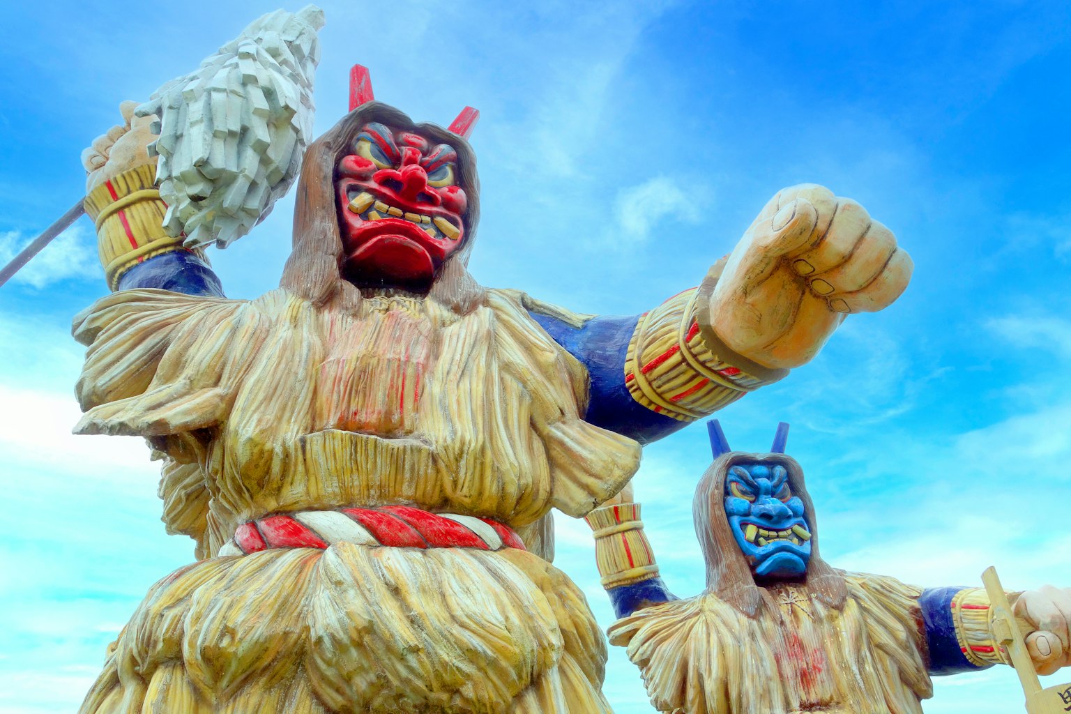 Zwei große Dämonenstatuen stehen unter einem blauen Himmel mit roten und blauen Gesichtern in strohähnlichen Kostümen