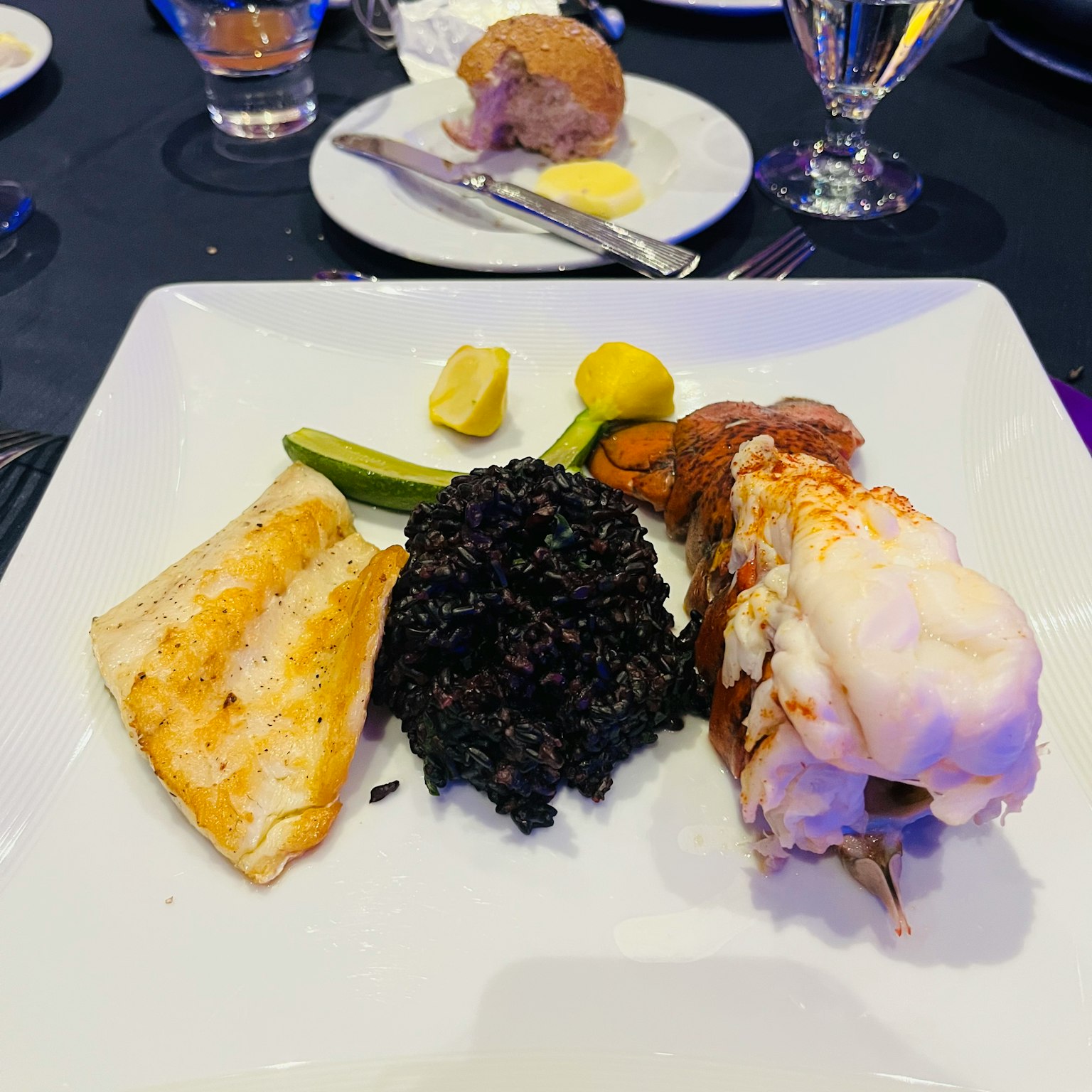 Seafood dish on a white plate featuring lobster black rice white fish green vegetables and lemon slices