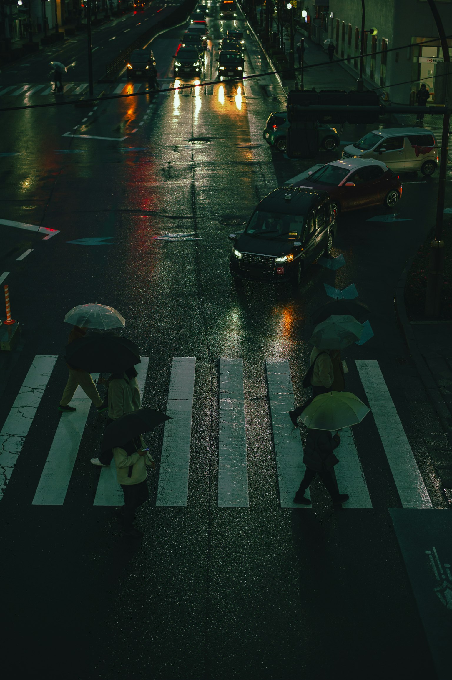 Night city scene with people crossing a crosswalk under umbrellas and cars passing by in the rain