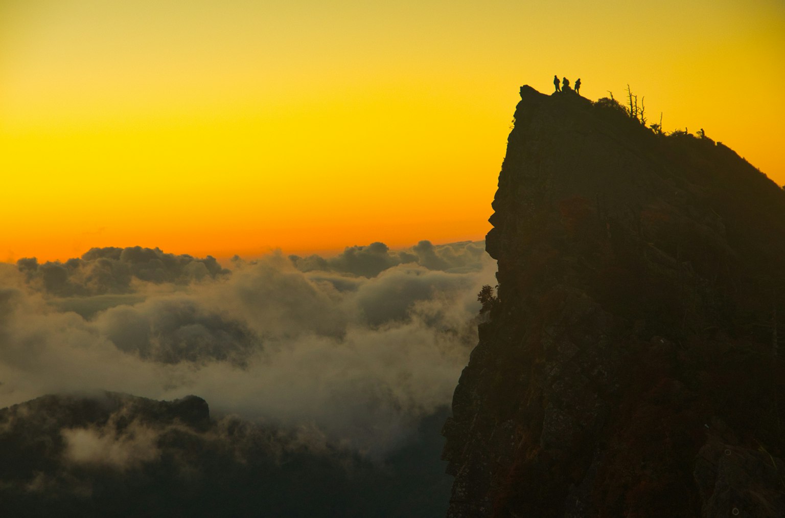 Silhouette di persone in piedi su una vetta montuosa al tramonto con nuvole sottostanti