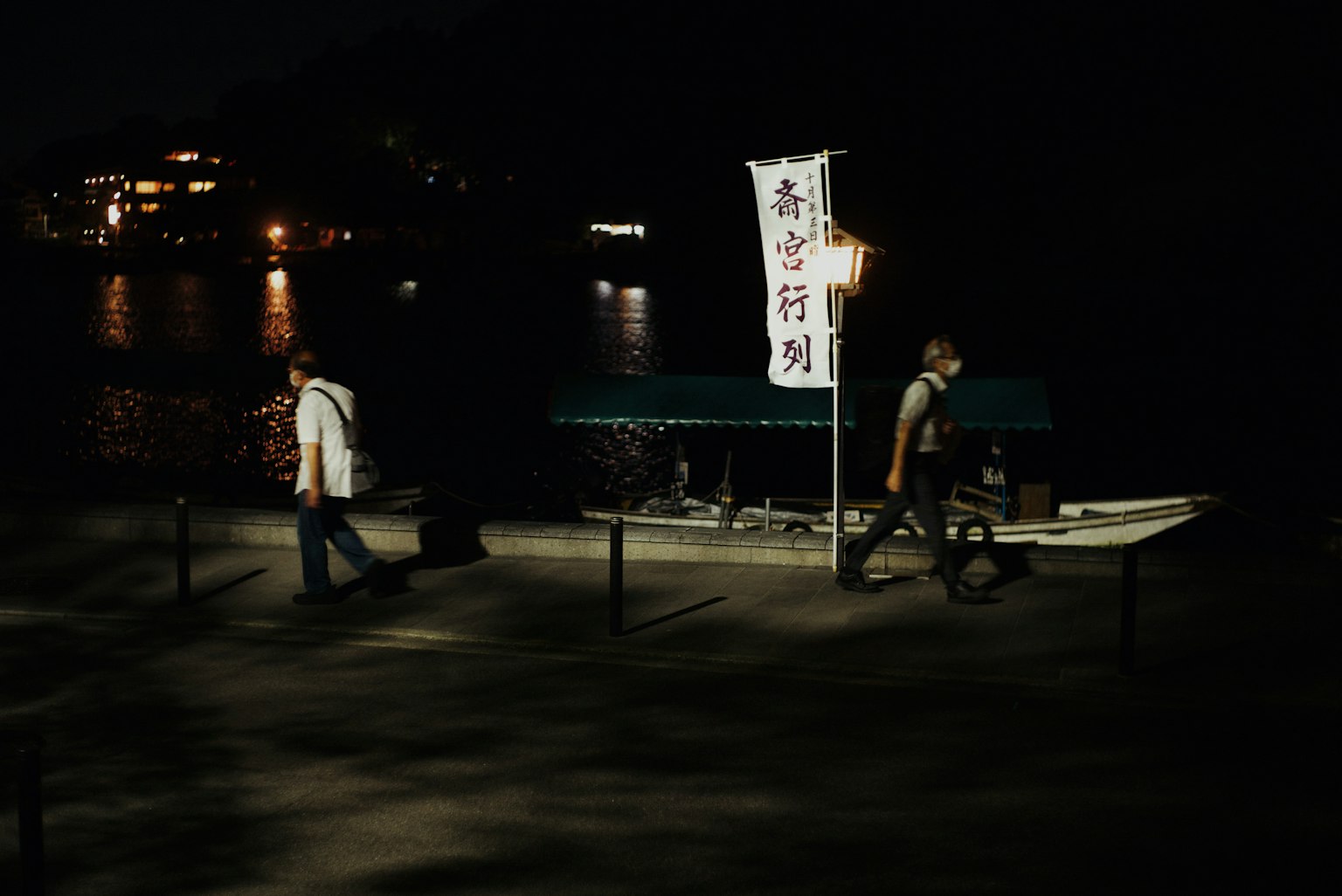 夜の川沿いを歩く二人の人物と小舟のある風景