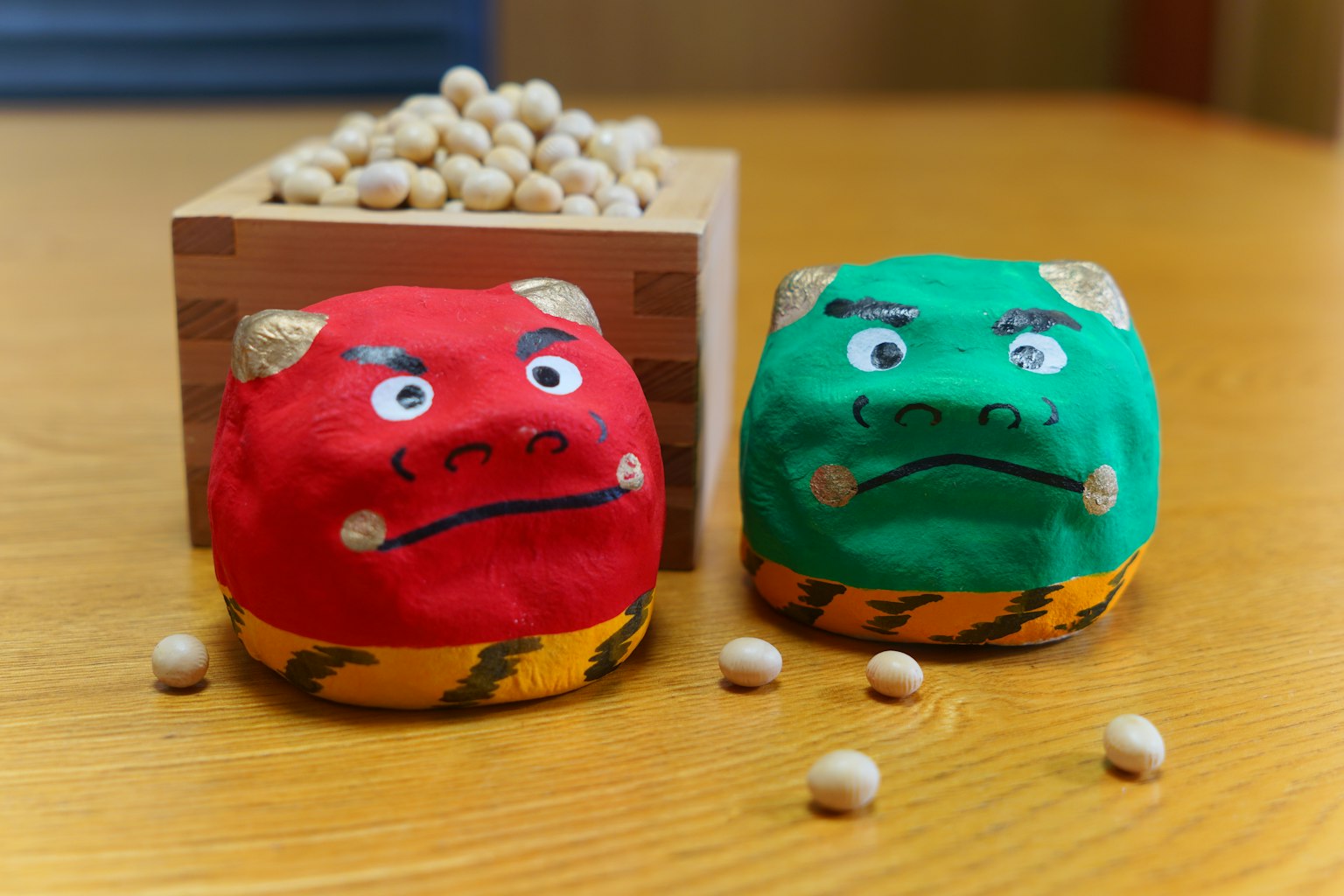 Figuras de demonios rojo y verde sobre una mesa con una caja de madera de frijoles al fondo