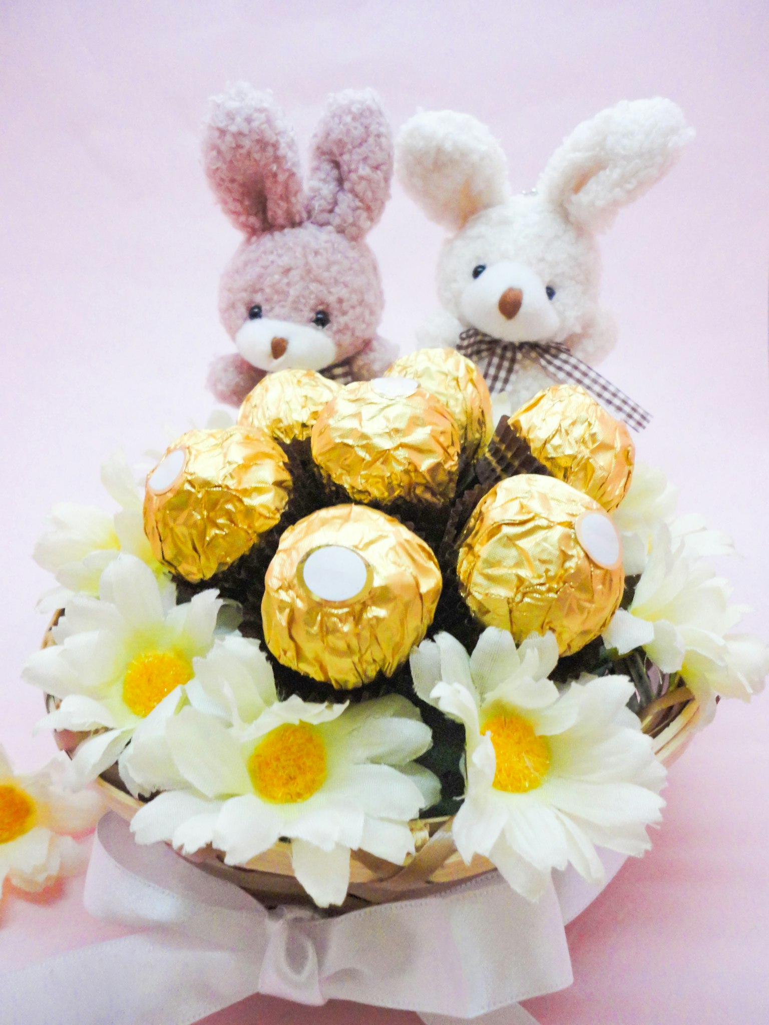 Una canasta con bolas de chocolate dorado y flores amarillas con dos juguetes de peluche de conejito sobre un fondo rosa