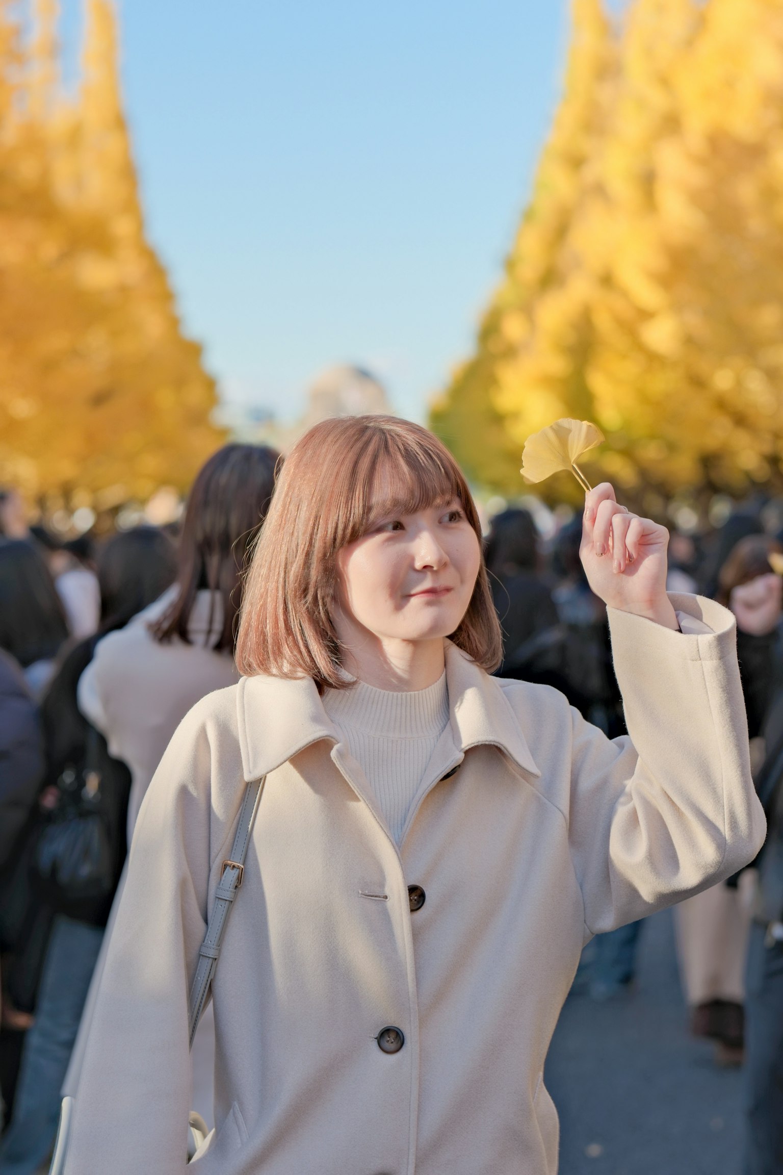 一位穿著白色外套的微笑女性在黃色銀杏樹下揮手