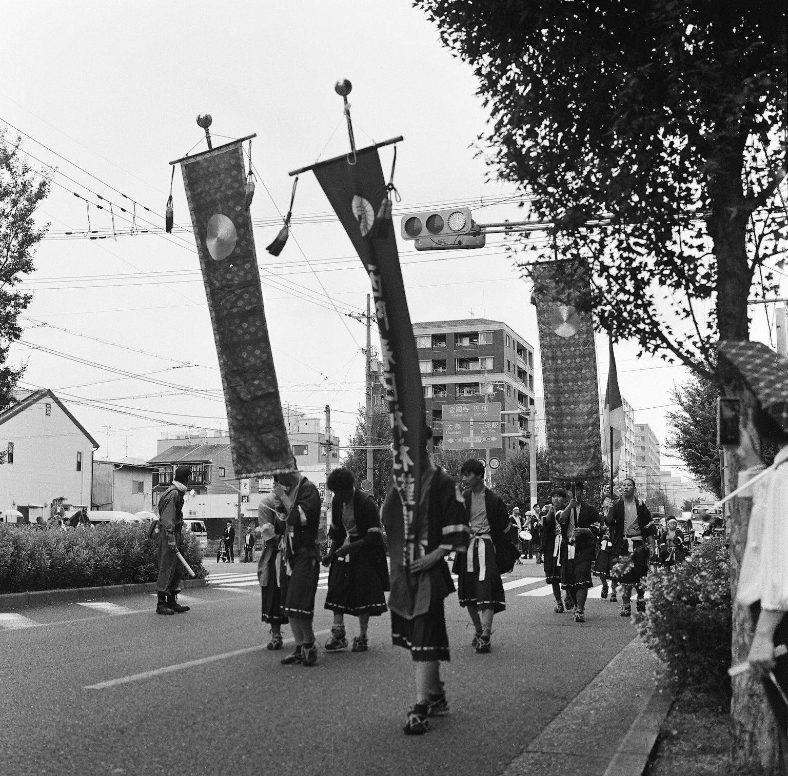 Schwarzweißfoto einer Festivalprozession, die die Straße entlanggeht, mit Teilnehmern, die große Banner halten