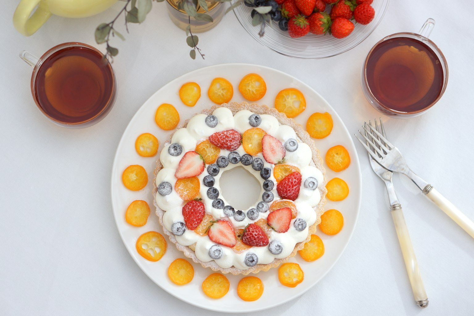 Kue buah yang indah disusun di piring putih dengan buah berwarna-warni dan dua cangkir teh di sampingnya