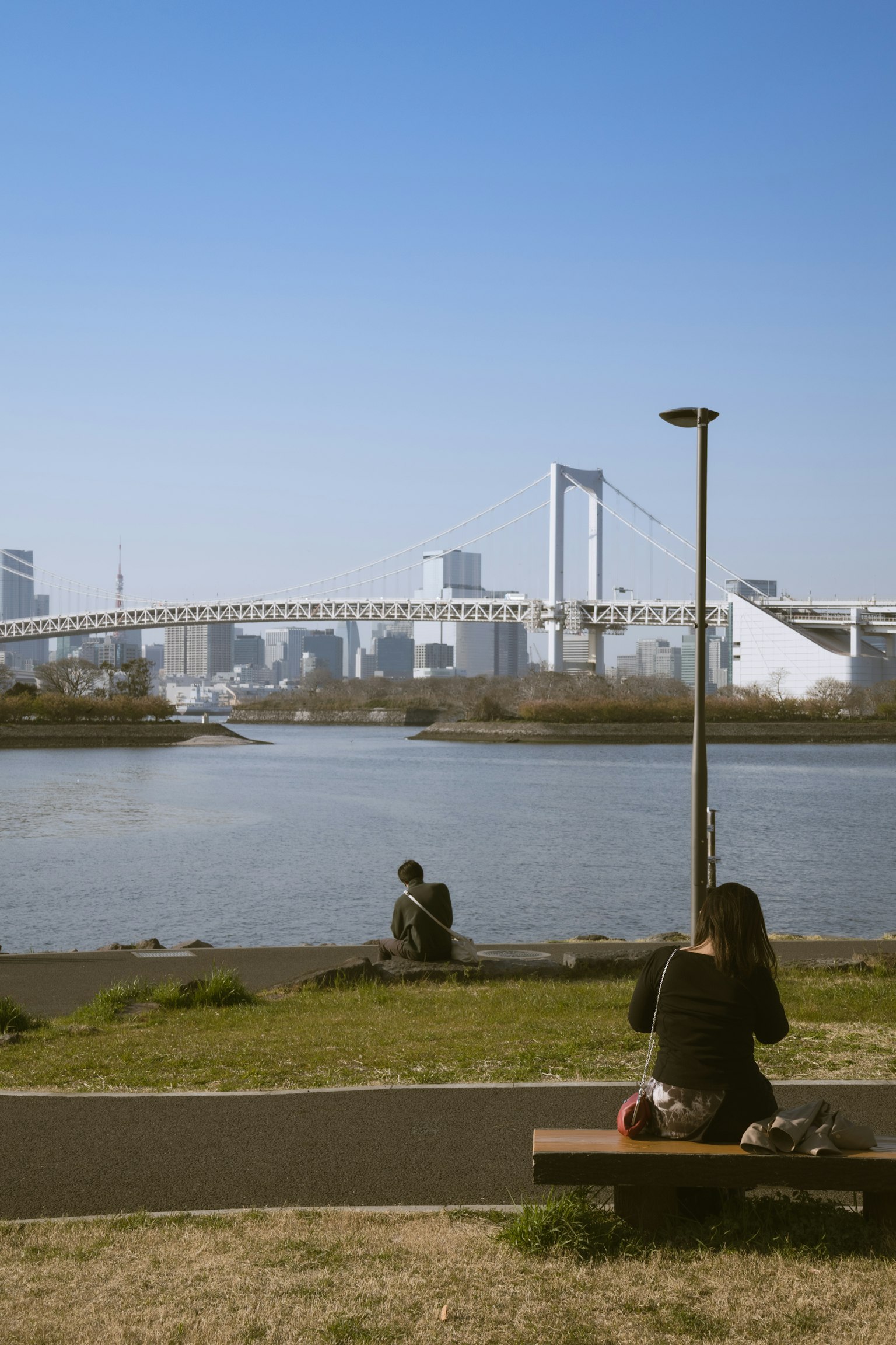 People relaxing near a river with a bridge and city skyline in the background clear blue sky