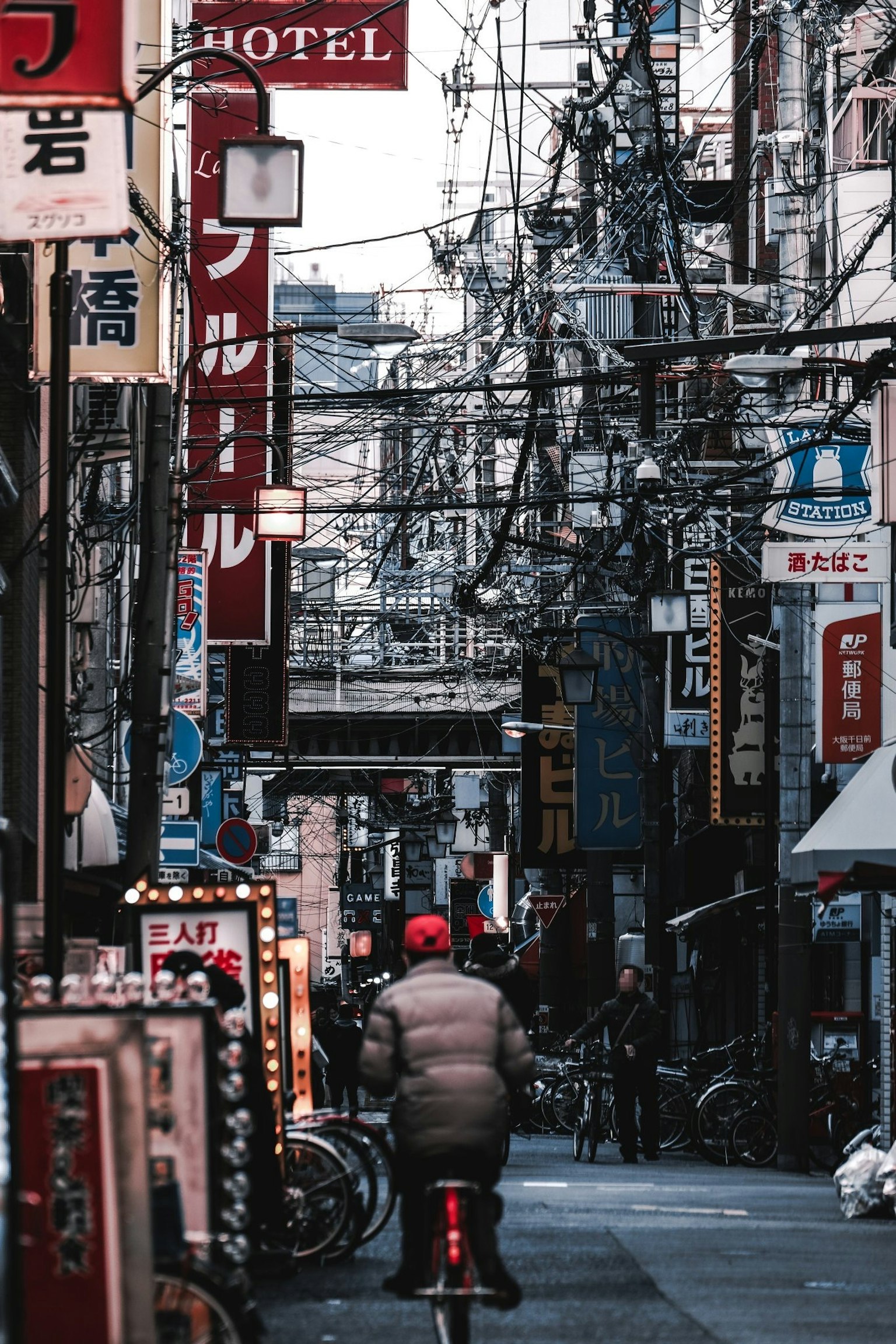 A narrow street scene in Tokyo with a person on a bicycle featuring tangled wires and various signs