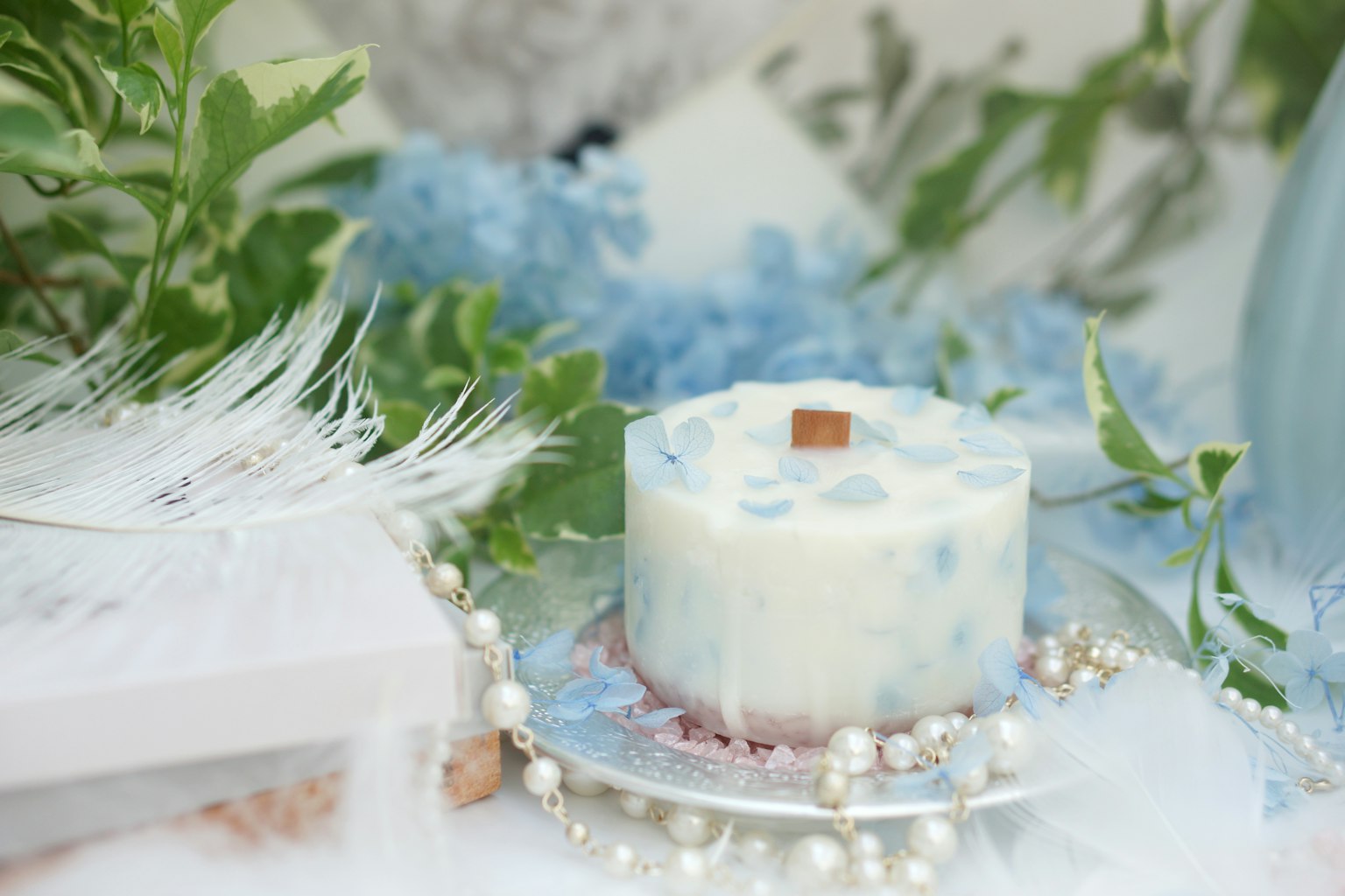 A scented candle with blue accents on a decorative plate surrounded by greenery and pearls