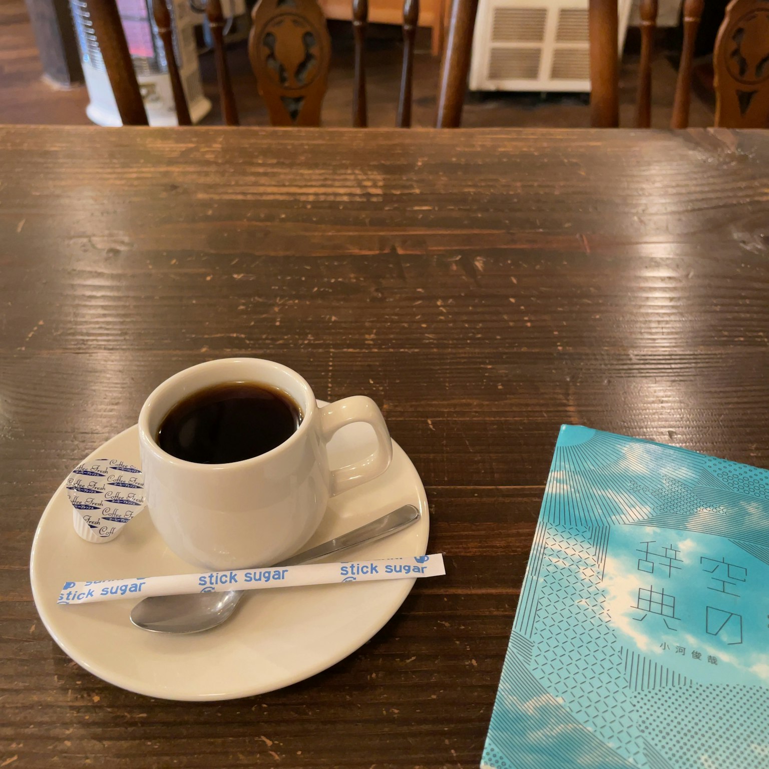 Coffee cup on a saucer with a spoon on a wooden table surrounded by wooden chairs and a blue book