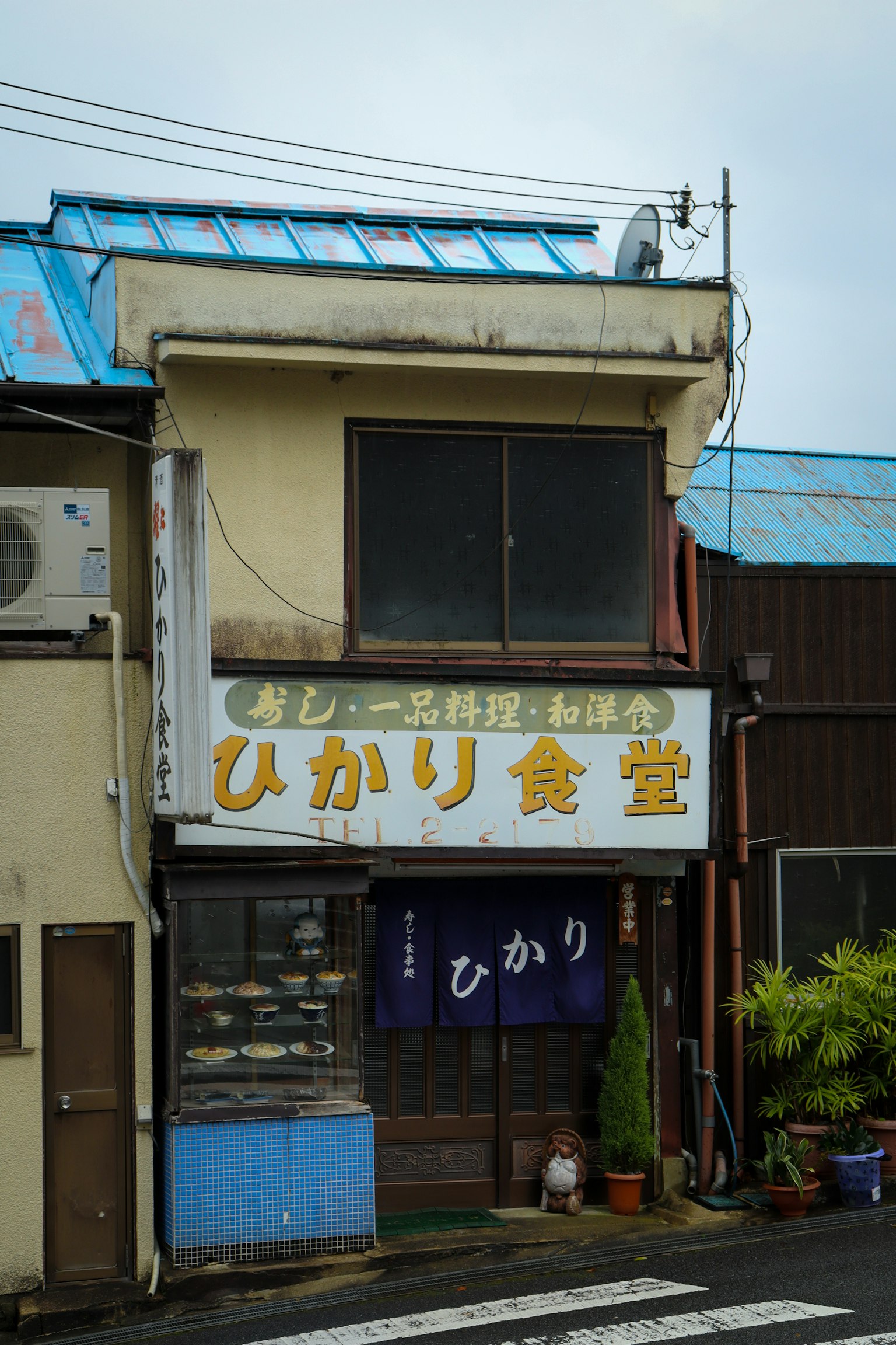 Exterior of Hikari Shokudo with blue roof and white sign visible plants and crosswalk