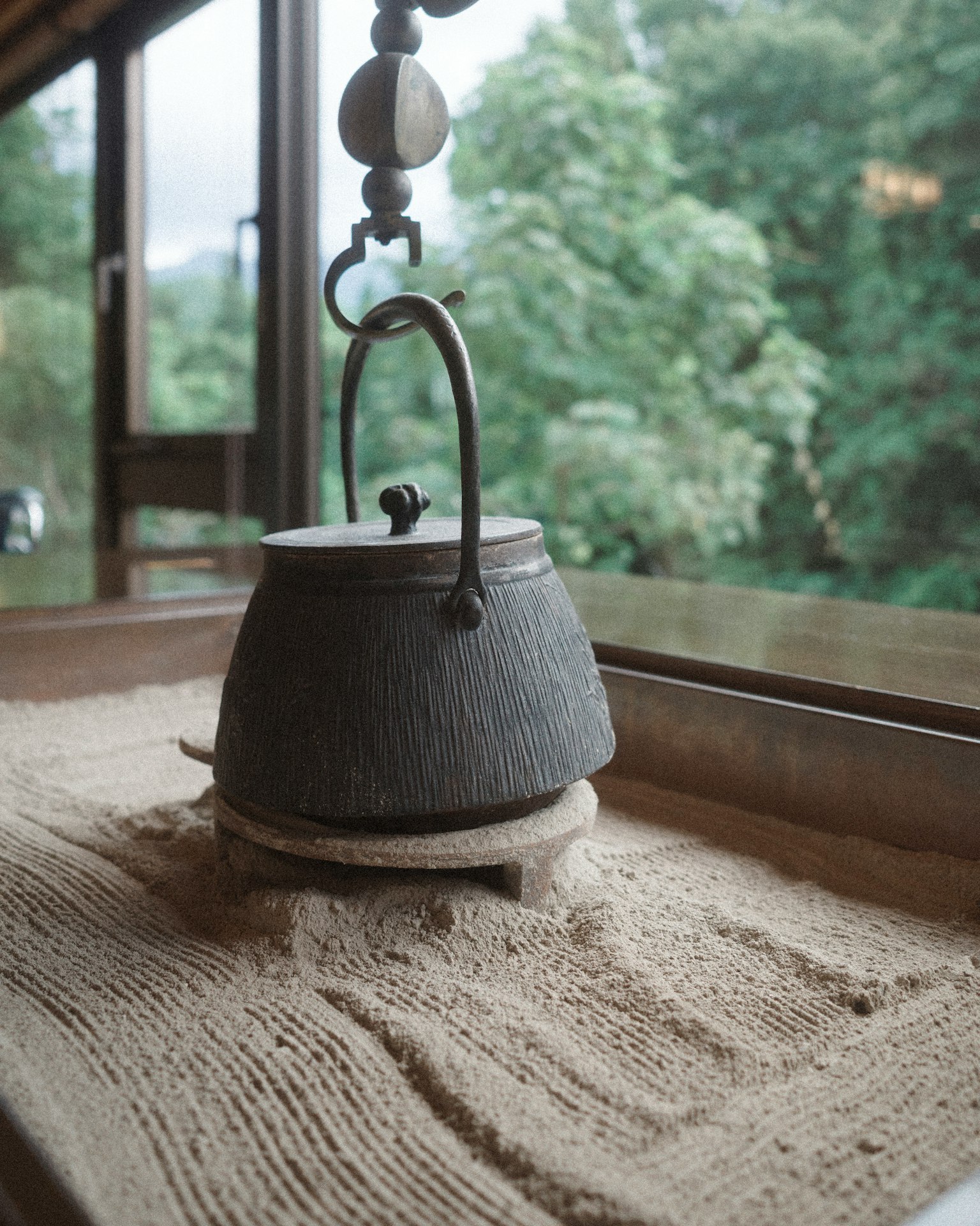 Une théière en fer noir posée sur du sable avec de la verdure visible à travers la fenêtre