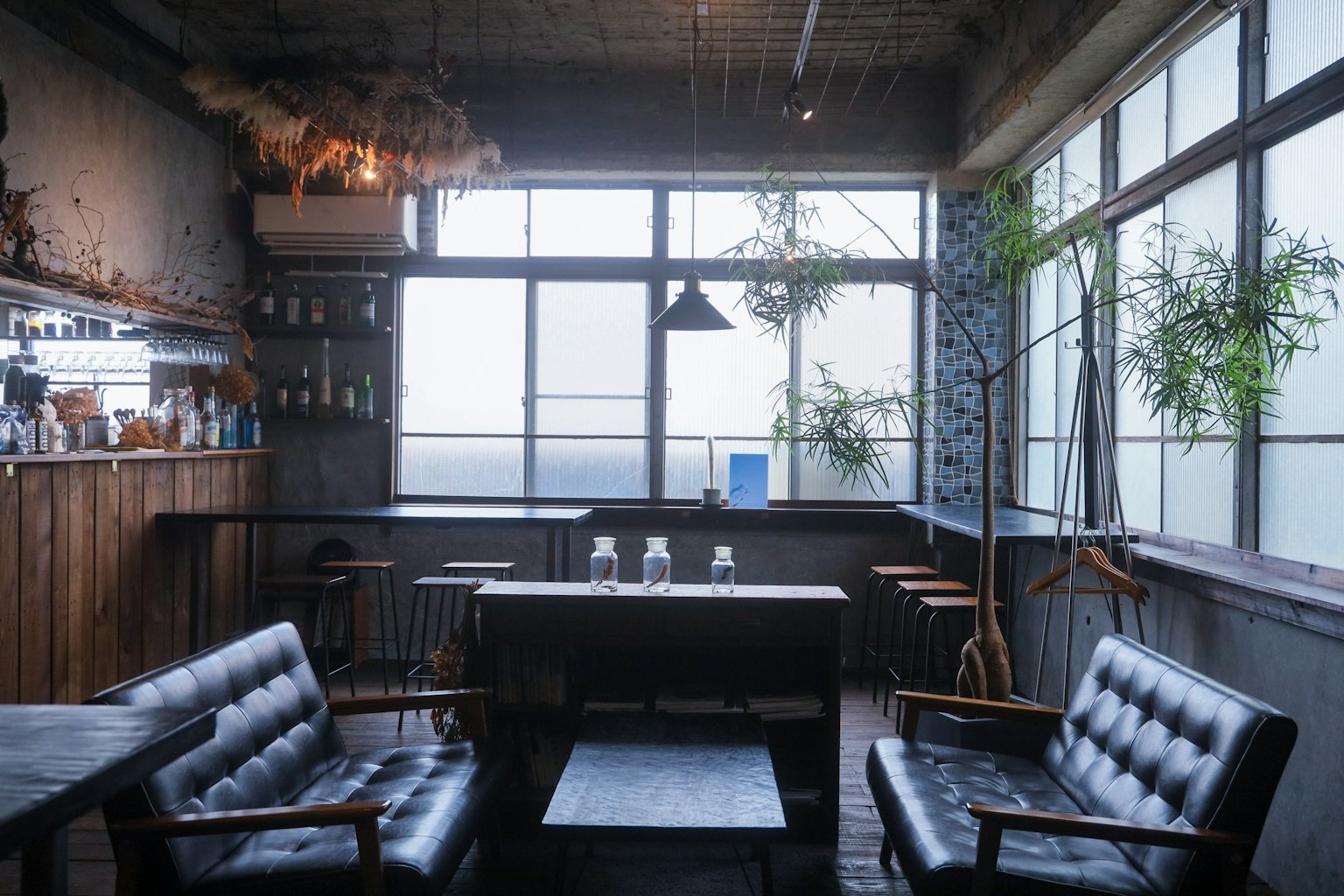 Interior de una cafetería con luz natural que entra por grandes ventanas con sofás negros y una mesa de madera