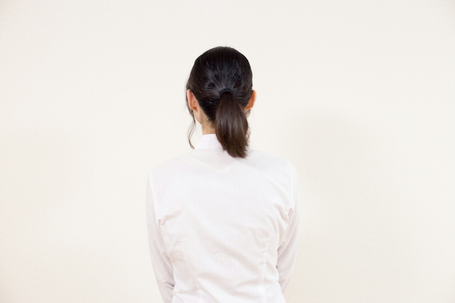 흰색 셔츠를 입은 여성이 단순한 배경 앞에서 뒷모습을 보이고 있다