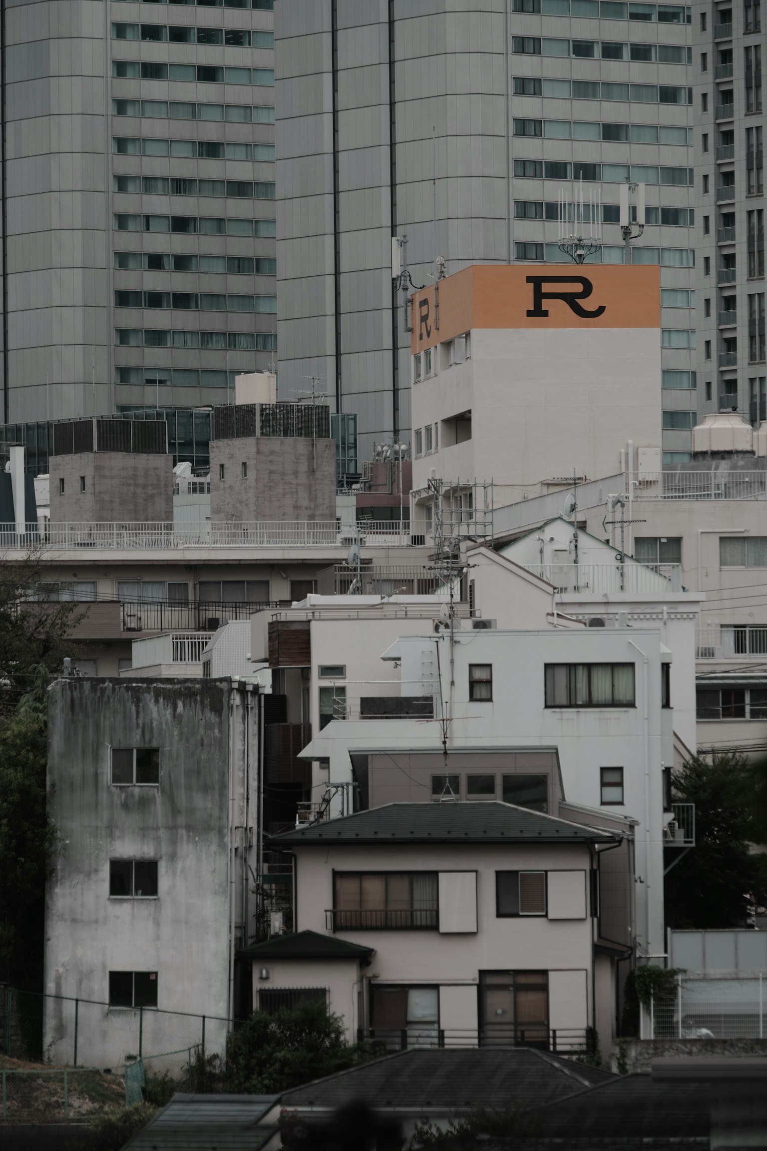Pemandangan kota dengan campuran gedung tinggi dan rumah tua, logo 'R' oranye yang mencolok