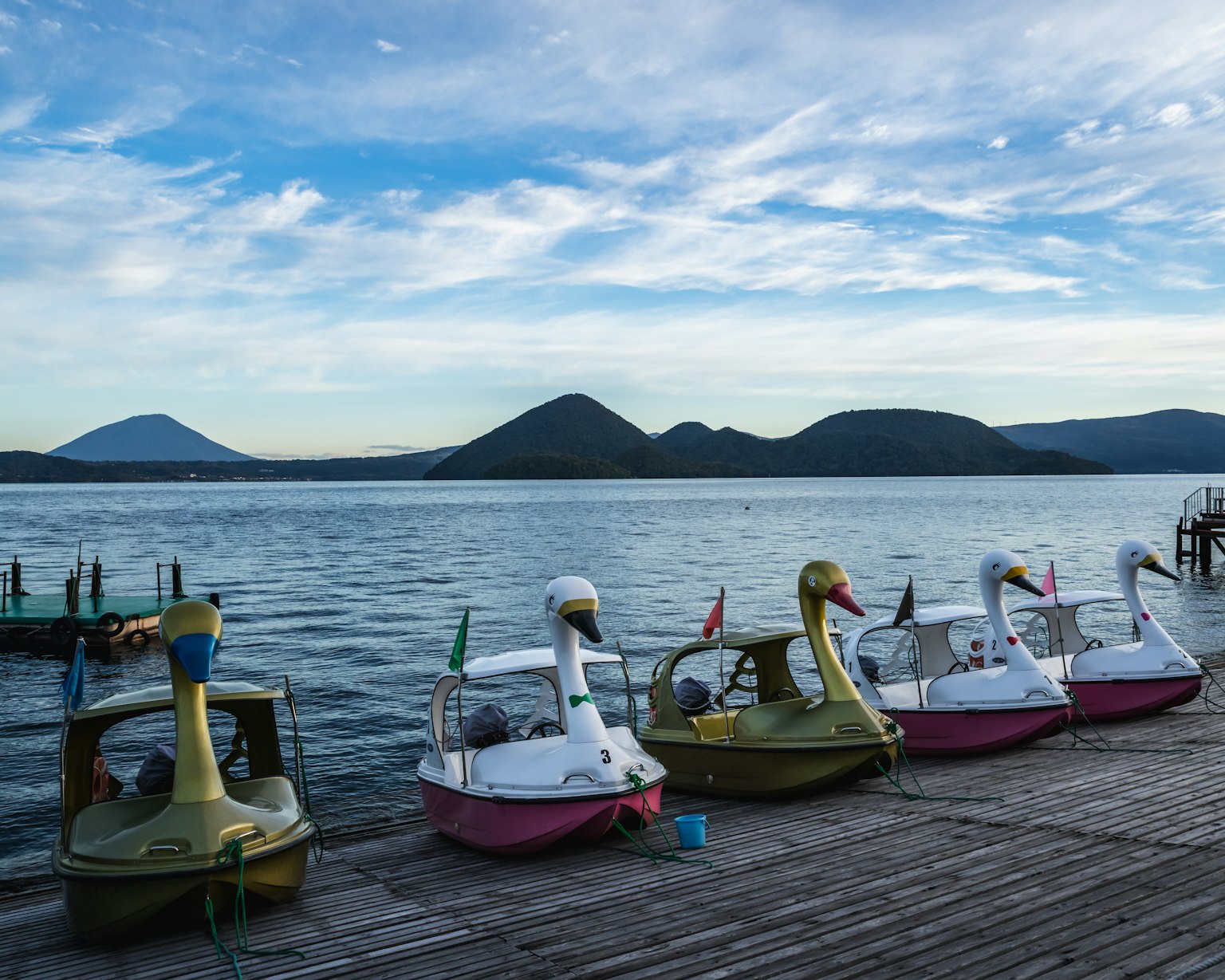 Fila de botes de pedal en forma de cisne en un lago con montañas al fondo y cielo azul