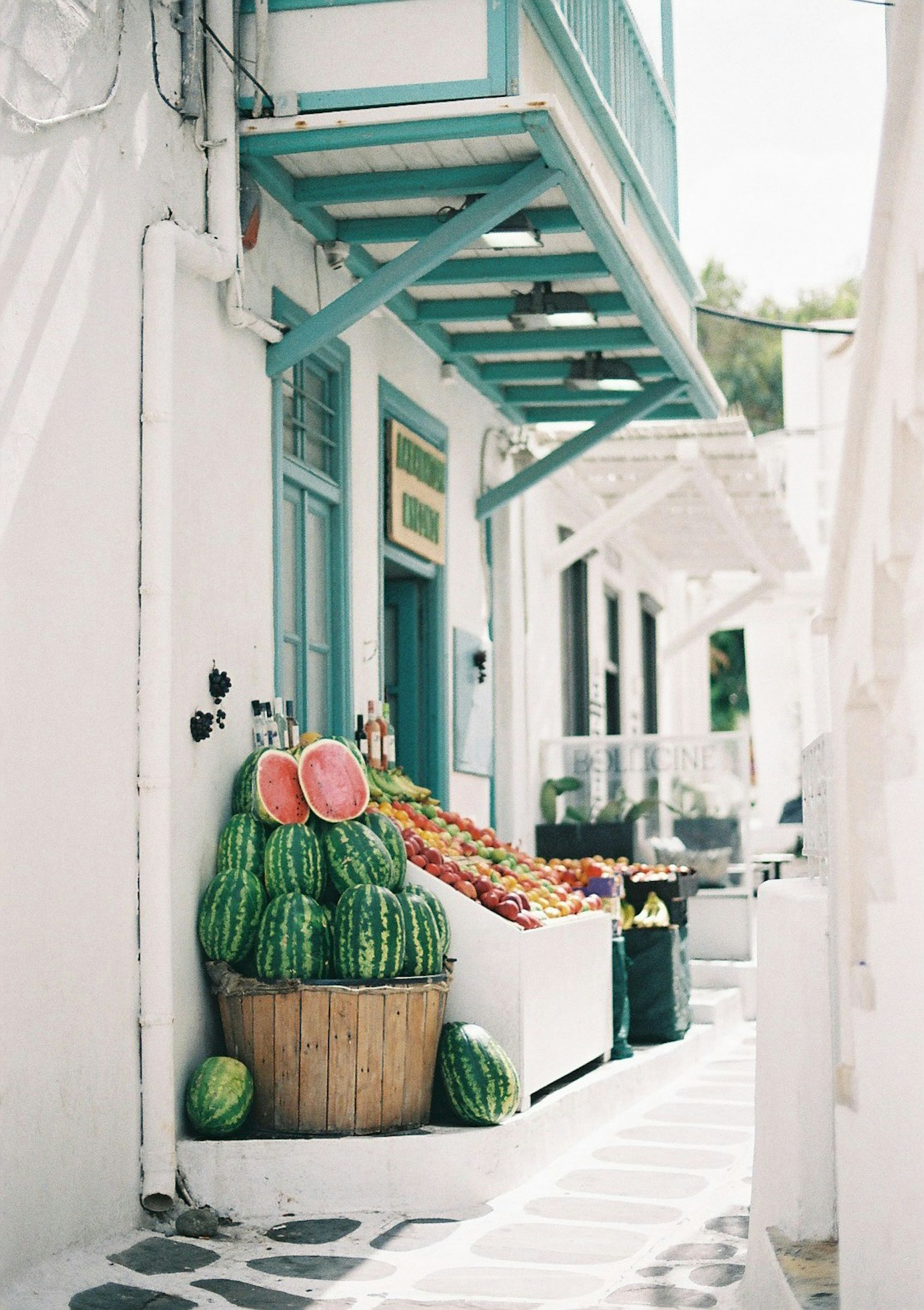 A charming Greek street scene featuring a basket of watermelons and various fruits against white walls