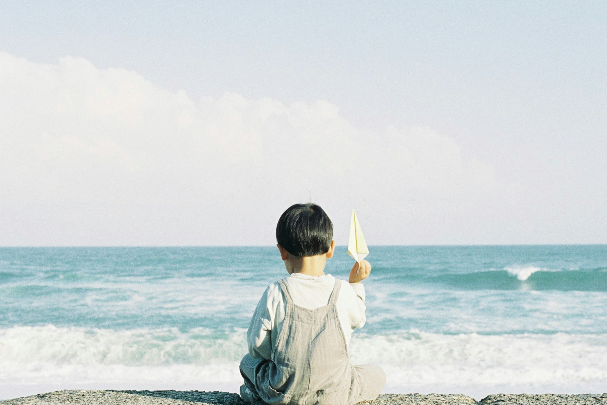 面向大海的孩子背影，遠處有白色船帆的帆船
