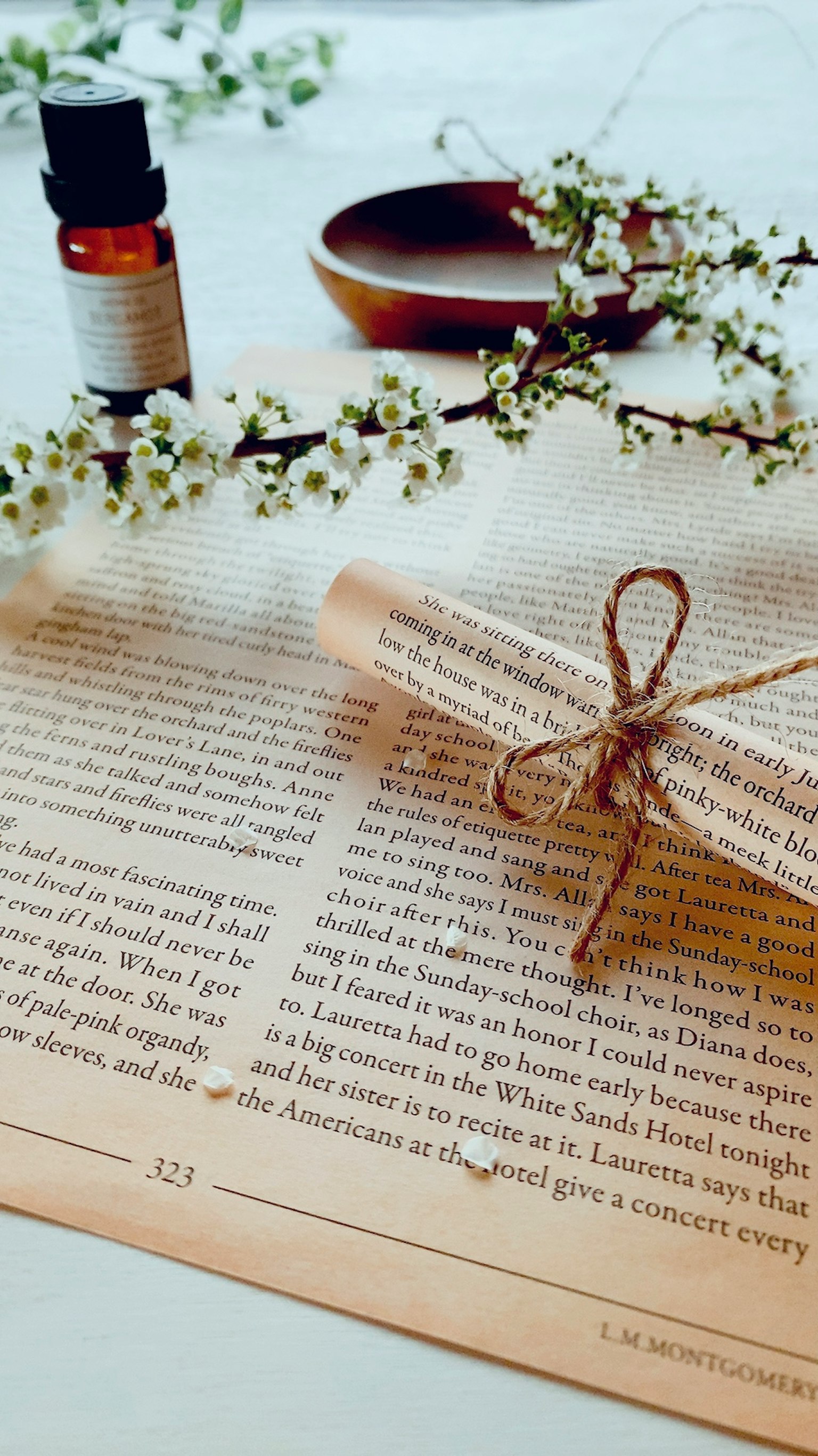 A scroll tied with twine on a book page accompanied by an essential oil bottle and floral branches