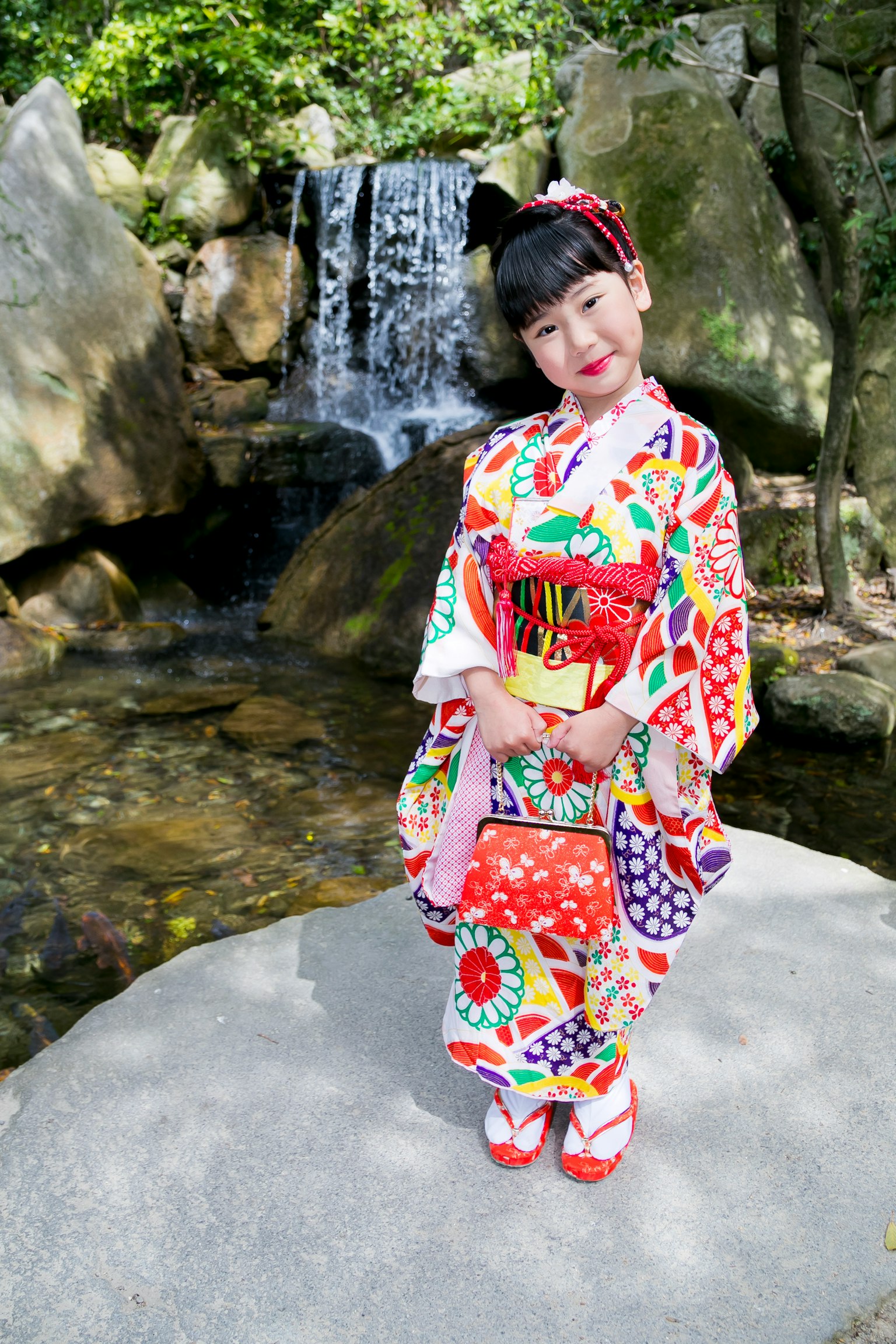 Girl in colorful kimono standing in front of a waterfall Bright floral patterns Red hair accessory Natural scenery