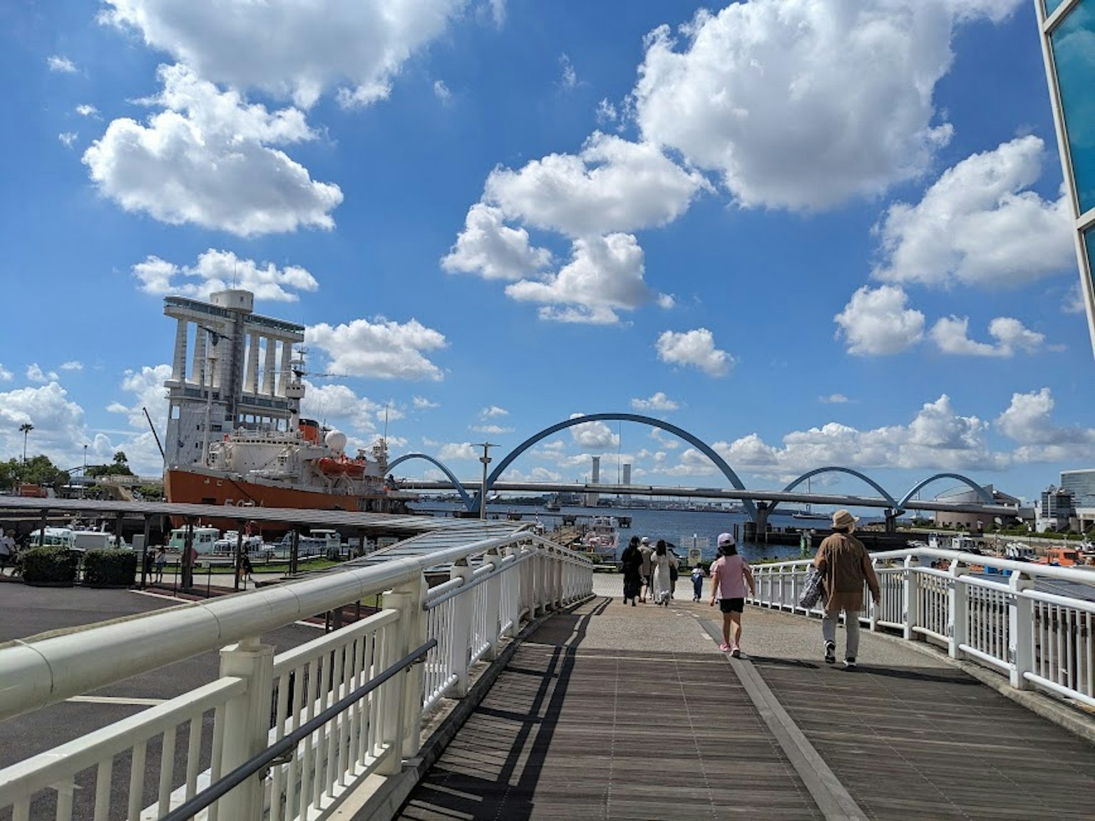 People walking along a boardwalk by a harbor with a ship and blue skies