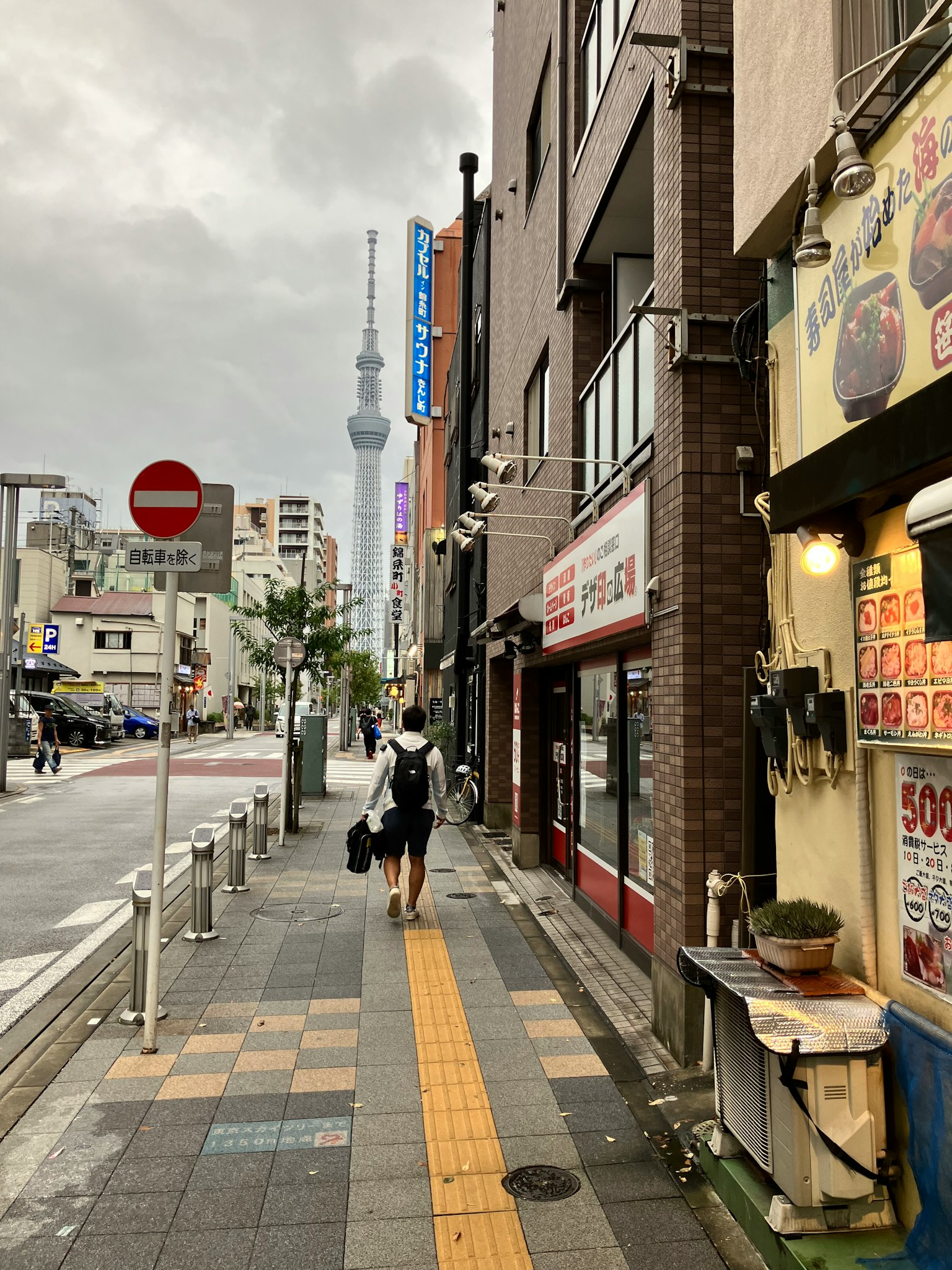 Una persona che cammina lungo una strada con la Tokyo Skytree visibile sullo sfondo sotto un cielo nuvoloso