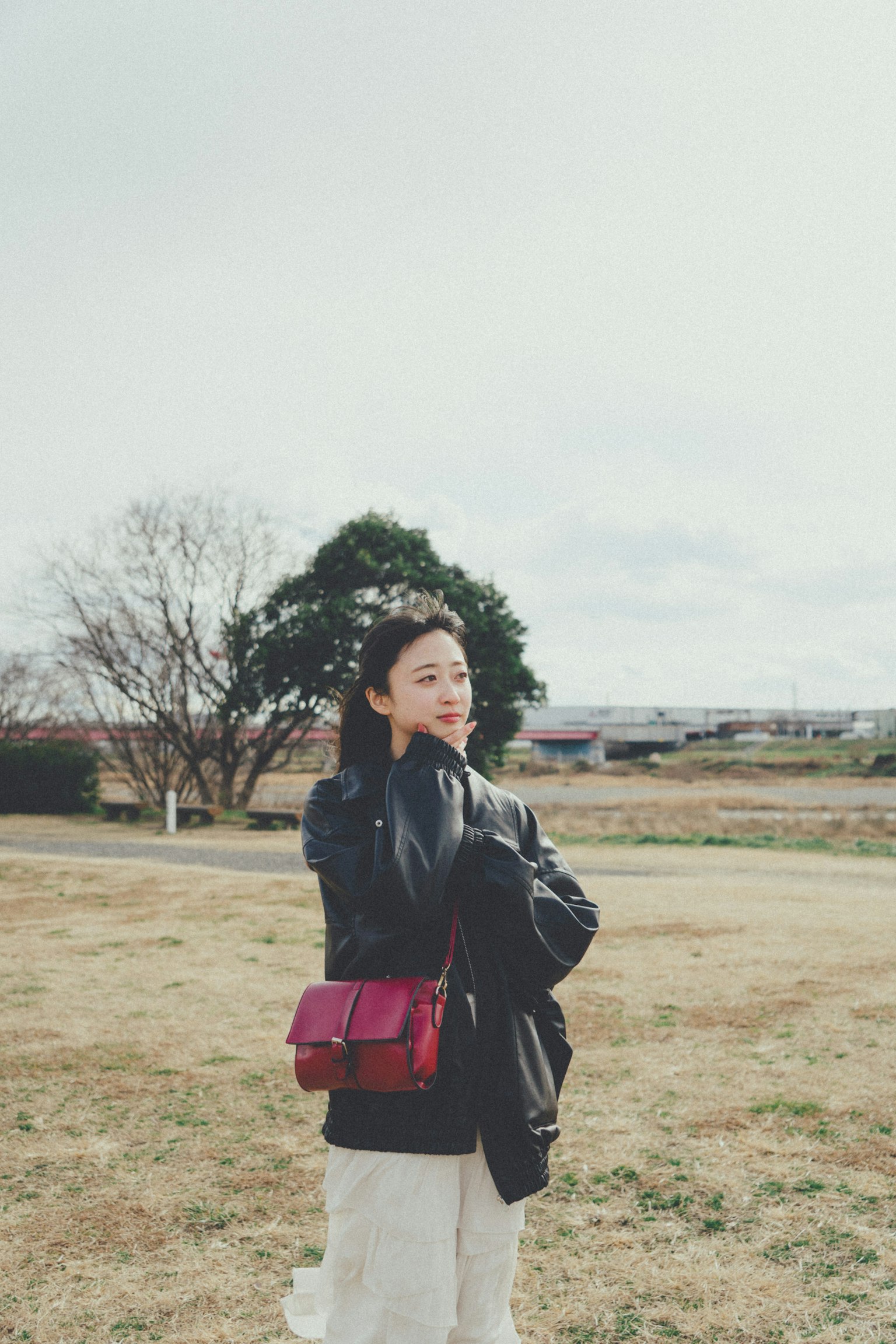 Una mujer posando en un parque con una chaqueta negra y pantalones blancos sosteniendo un bolso rojo