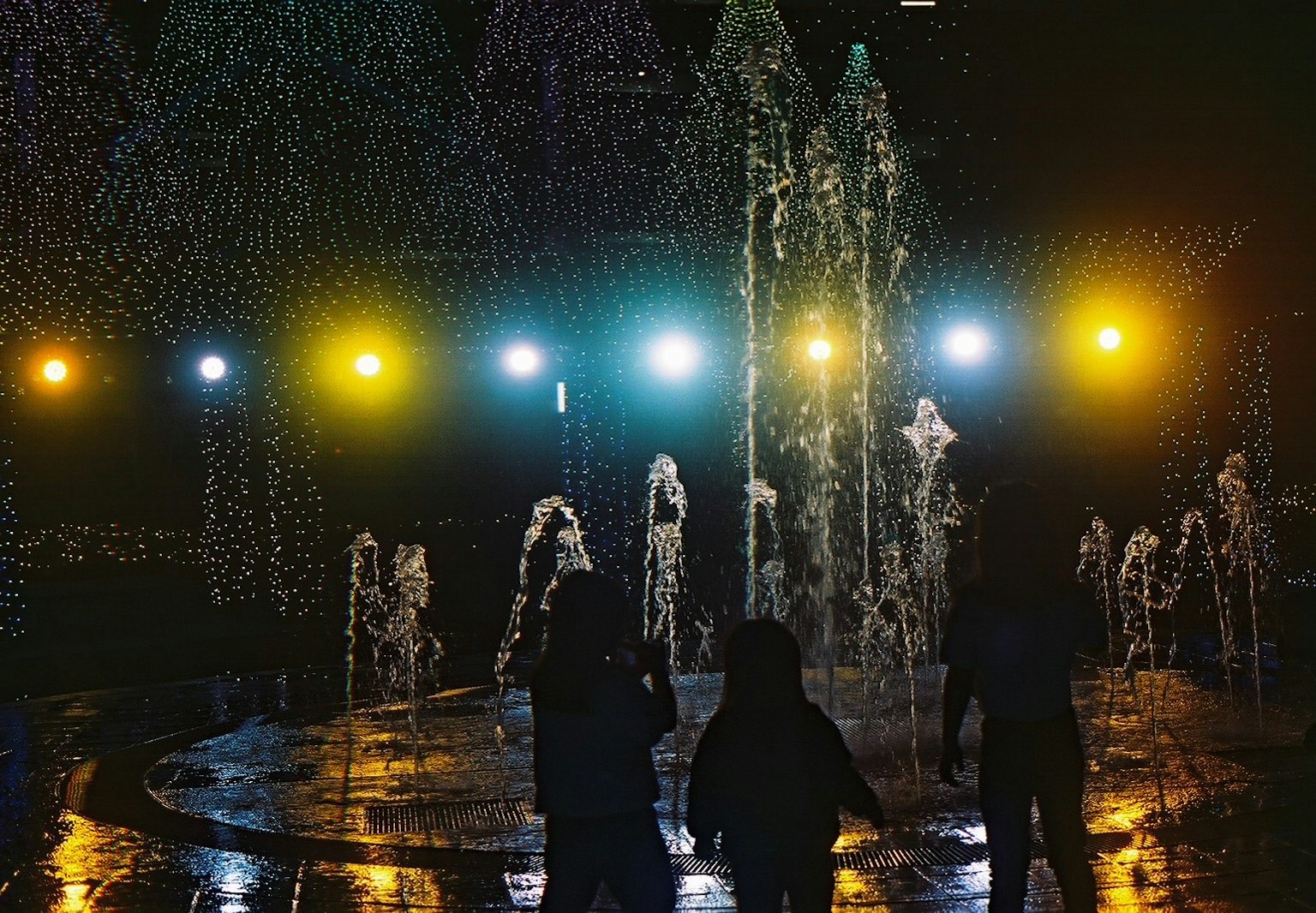 Silhouetten von Menschen vor einem beleuchteten Brunnen bei Nacht