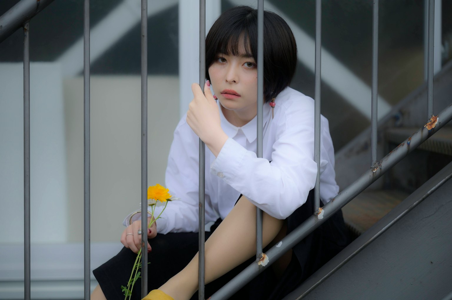 Una estudiante mirando a través de barandillas metálicas en una escalera vestida con una camisa blanca y una falda negra sosteniendo una flor amarilla
