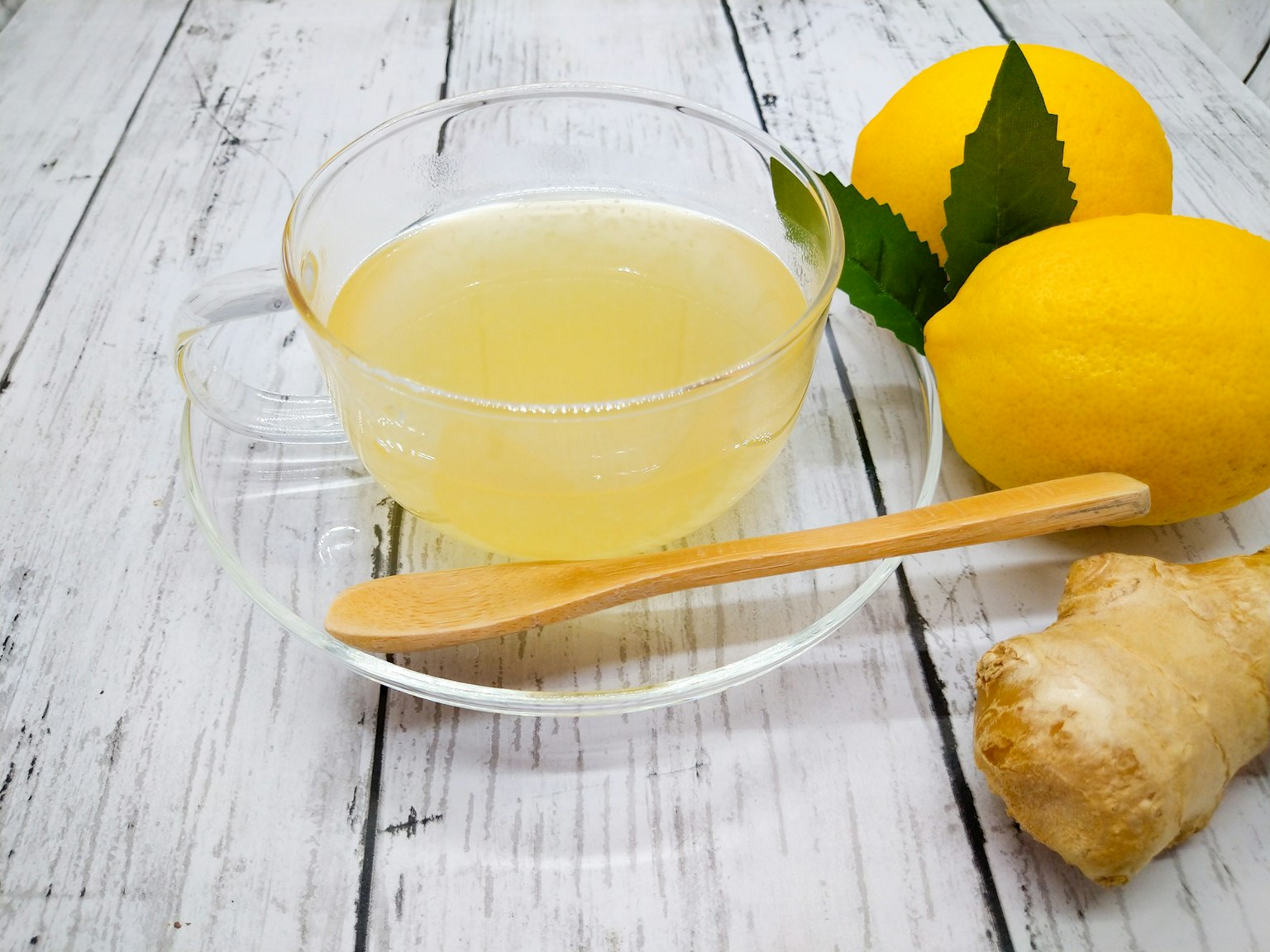 Secangkir minuman lemon jahe bening dengan lemon segar dan akar jahe di dekatnya