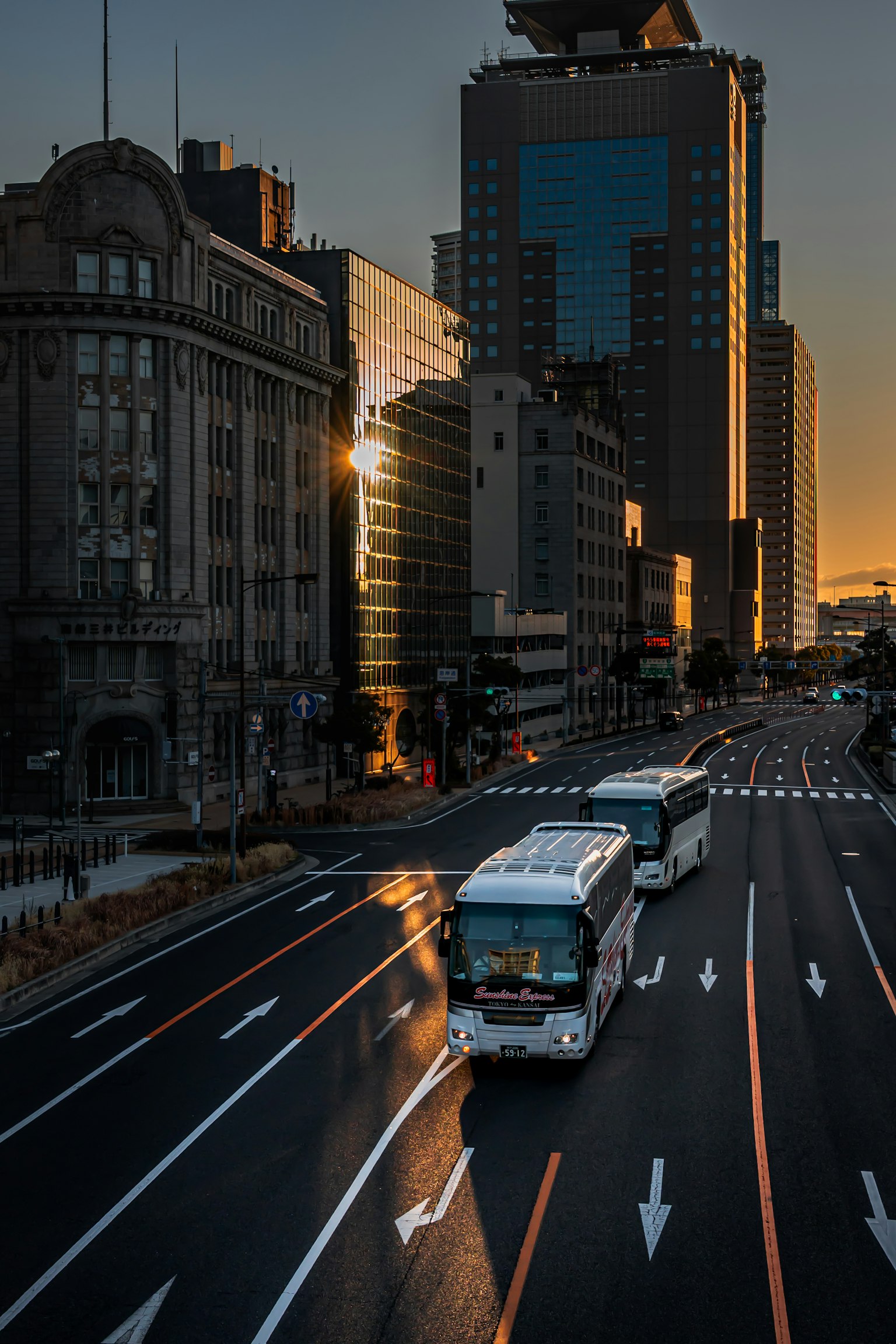 Buses driving along a city road at dusk with surrounding buildings