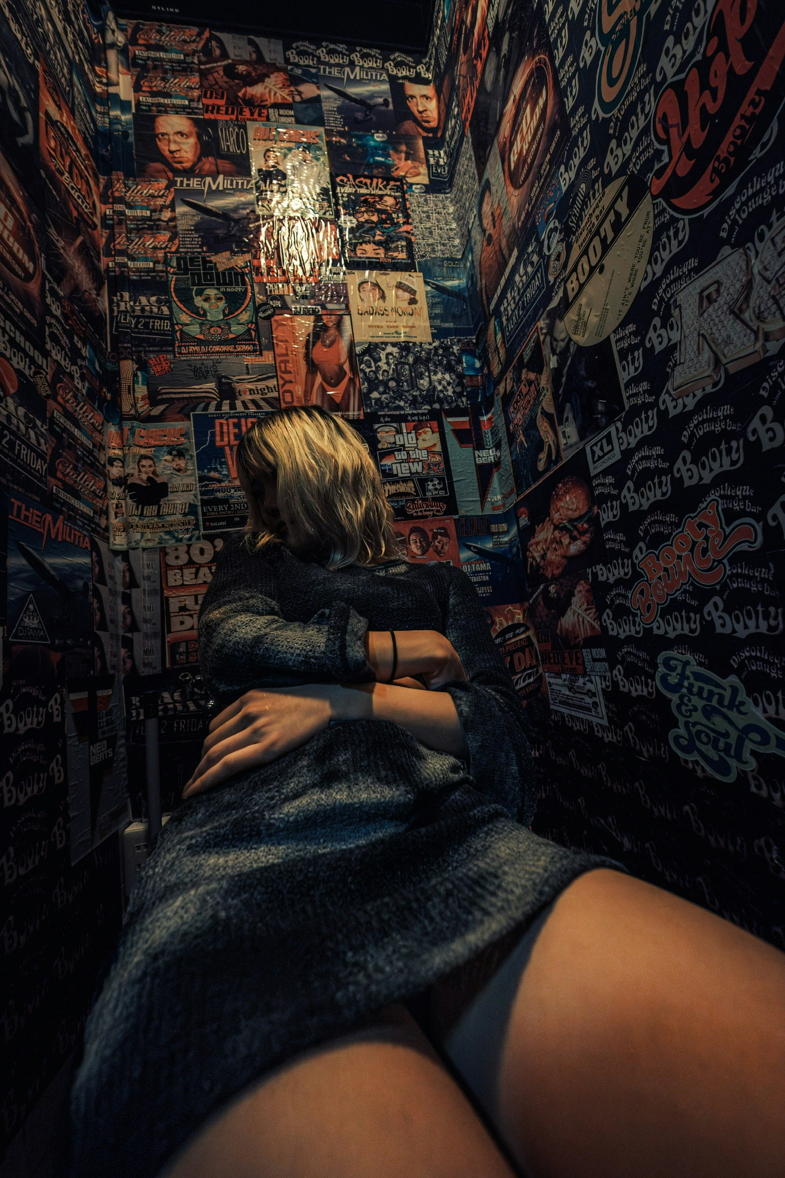 Seorang wanita duduk di toilet sempit dengan grafiti berwarna dan poster di dinding