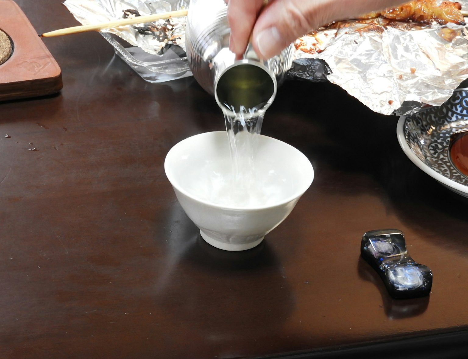 手でお酒が注がれている白い茶碗とテーブルの上にある料理