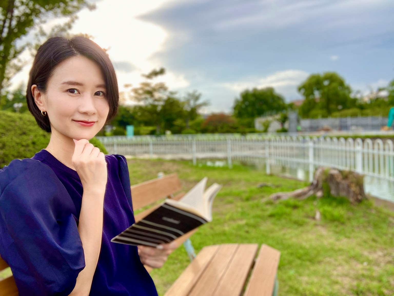 一位女性坐在公園長椅上閱讀一本書 穿著藍色襯衫 拿著一本黑色書籍 周圍是綠色樹木和寬闊的天空