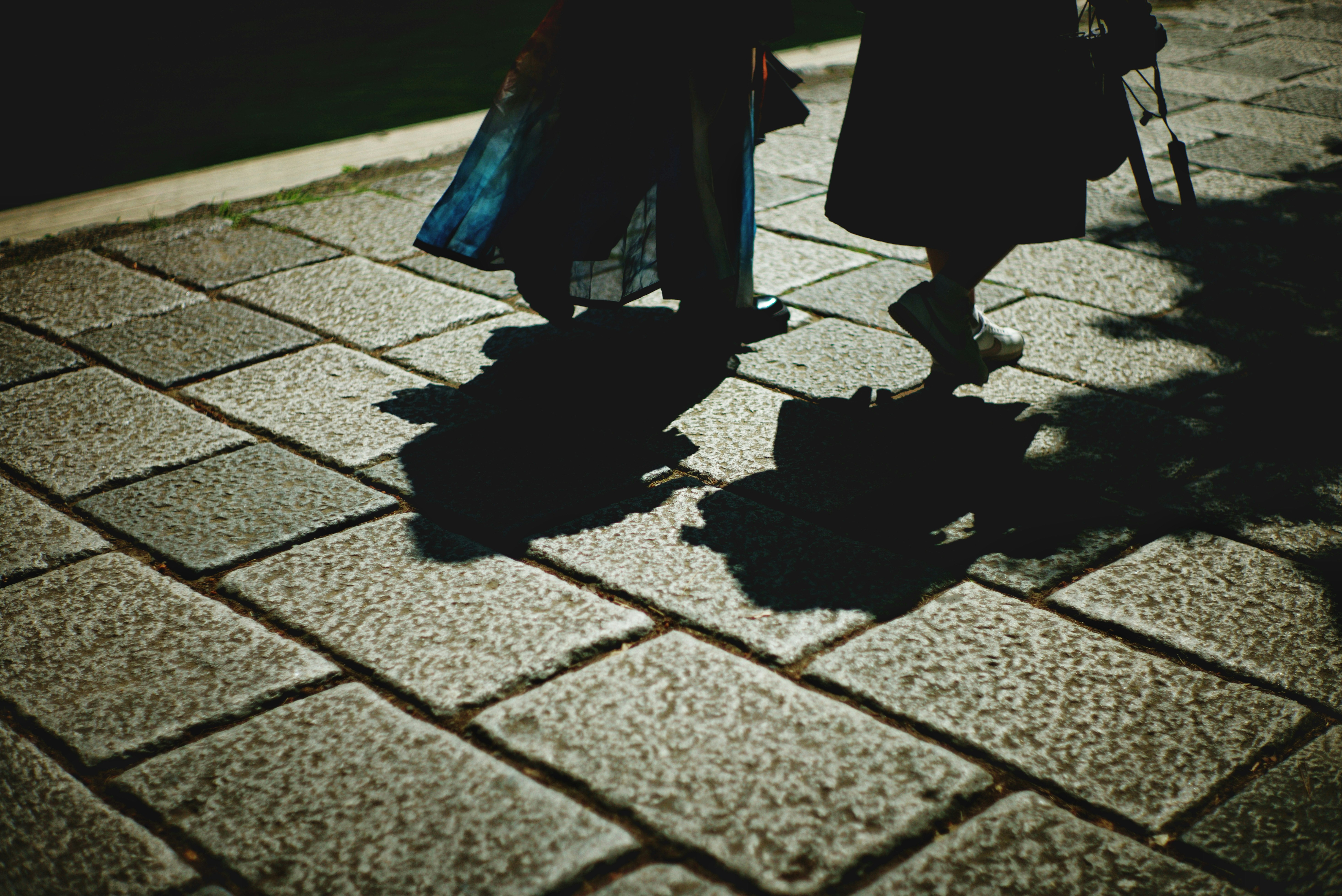 Two figures walking on a stone pavement casting shadows