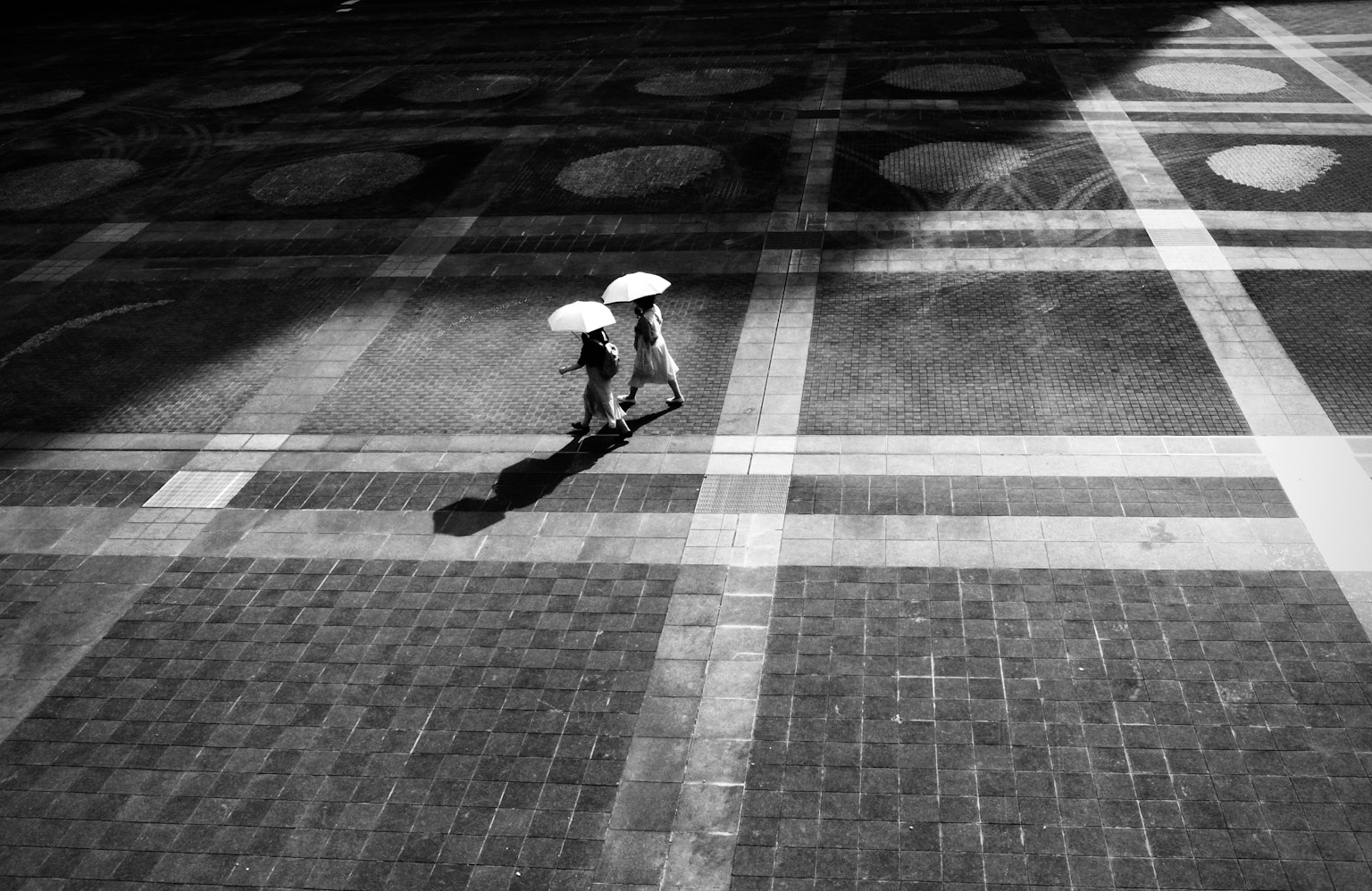 Black and white street scene with two people walking on tiled pavement holding umbrellas