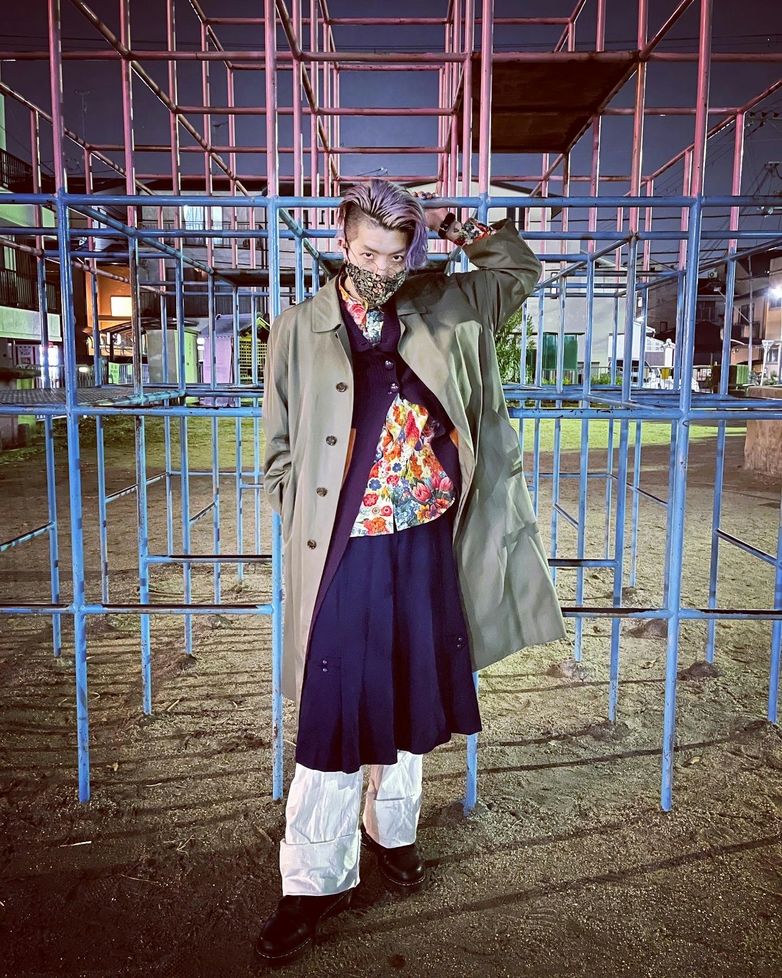 Una persona in abiti colorati si trova in un parco notturno con una struttura in acciaio blu sullo sfondo