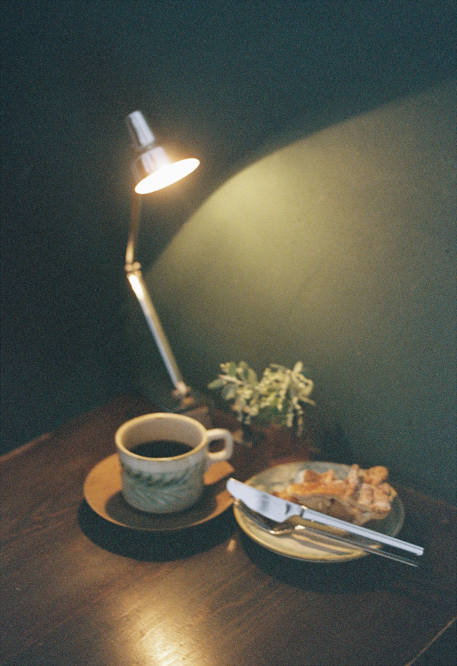 Coffee cup on a wooden table with a small plant and a plate of food beside a warm light lamp