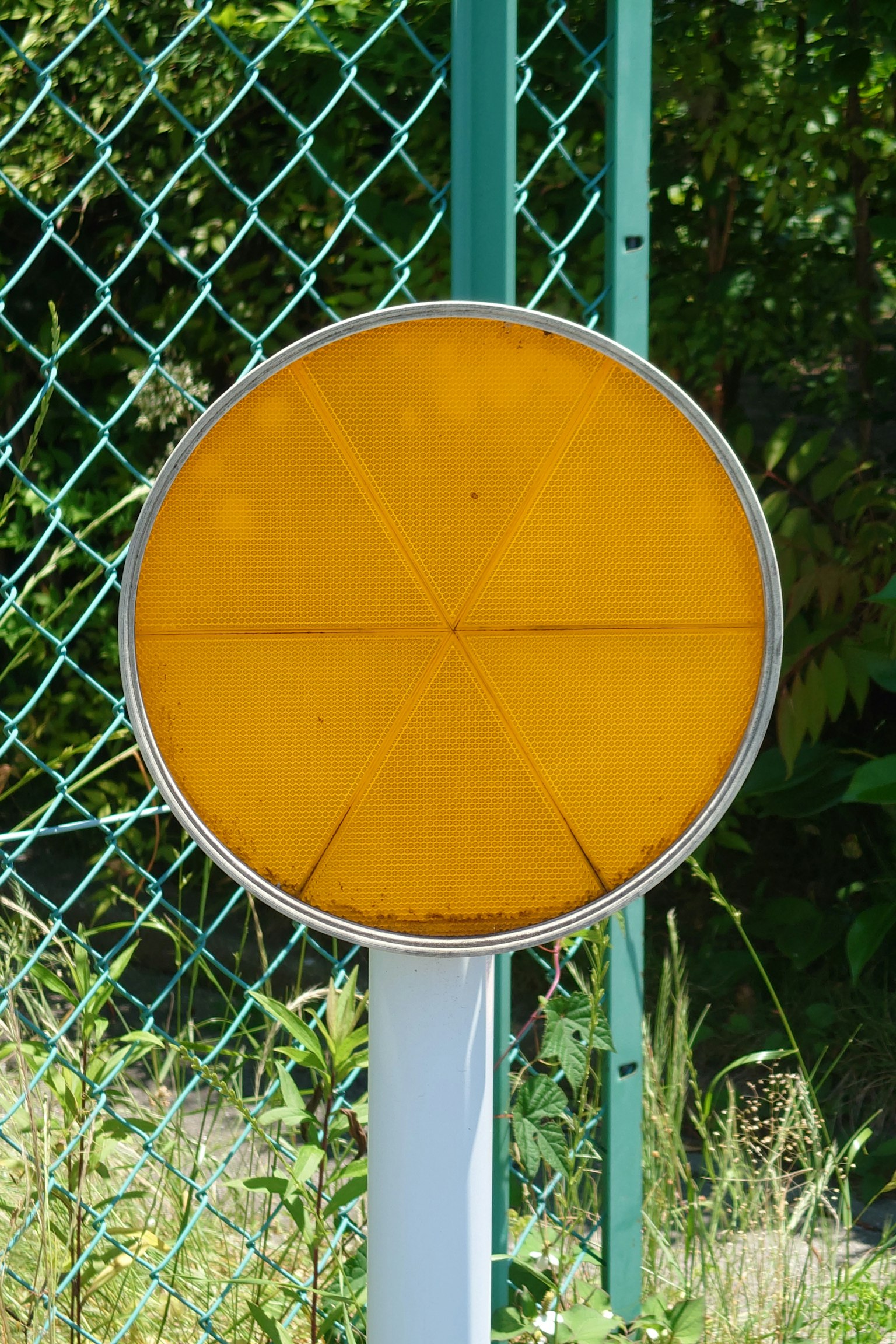 Un panneau circulaire jaune se dresse devant une clôture verte